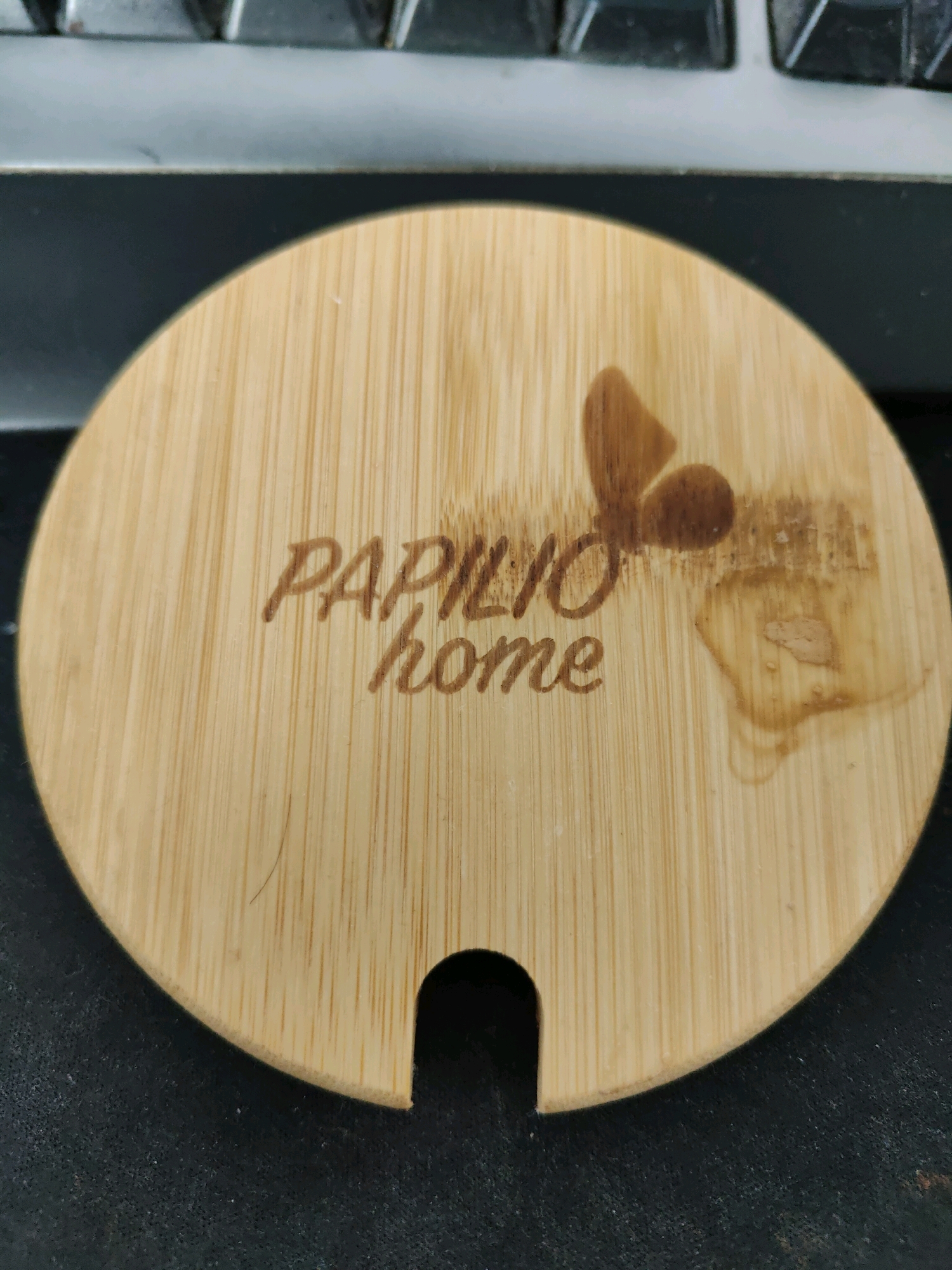 This image shows a close-up, overhead view of a round, light-colored bamboo lid with a small crescent-shaped cutout on the bottom edge. The lid is engraved with the words "PAPILIO home" in a cursive font. To the left of the text, there is a brown, engraved image of a butterfly. The lid is resting on a black, textured surface, likely a desk or table. In the background, the edge of a keyboard is visible, with gray keys and a silver trim. The overall lighting suggests an indoor setting. The image does not contain any information about the city of Homyel, Belarus.