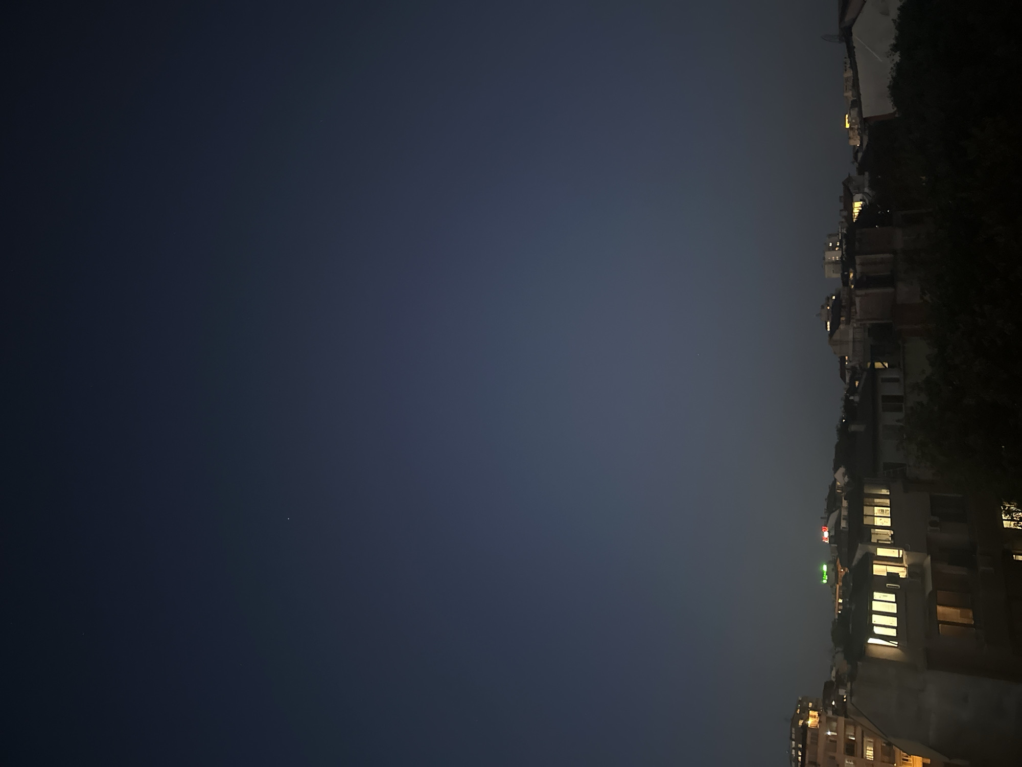 The image captures a cityscape at dusk, likely in Varna, Bulgaria, as indicated by the presence of the city's iconic Palace of Culture and Sports in the distance. The sky is a deep indigo, transitioning from the last vestiges of daylight. A few stars are faintly visible, adding to the serene atmosphere. Below, the silhouettes of buildings are illuminated by the warm glow of lights from windows, suggesting evening activity within. Some buildings are taller and more modern, while others appear older and more traditional. The foreground is dominated by the dark foliage of trees, partially obscuring the lower levels of some structures. The overall impression is one of a quiet urban evening, with the city settling into the night.