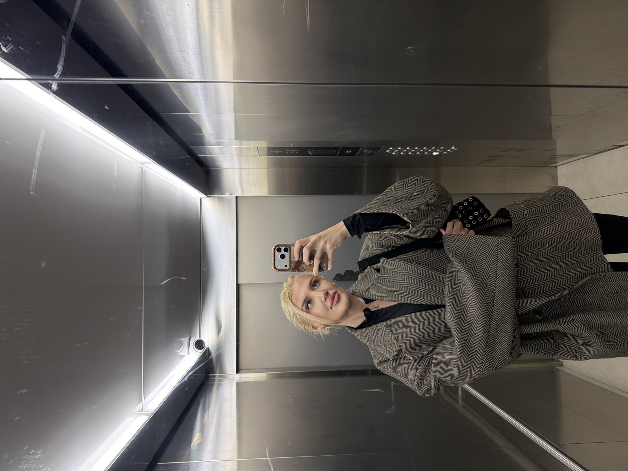 A blonde woman is captured in a selfie, taken from her reflection in the highly polished, metallic walls of an elevator. The original image is rotated clockwise by 90 degrees. She holds a smartphone with a light-colored, patterned case in her right hand, aiming it to photograph her reflection. Her gaze is directed towards the phone screen, with a neutral and composed expression.
She is dressed in a dark, structured coat or jacket featuring a distinctive herringbone pattern in shades of brown and black, worn over a black top. The elevator interior consists of sleek, reflective metal panels that create a mirrored effect throughout the space. Linear strip lighting runs along the ceiling edges, casting bright illumination and reflections across the shiny surfaces.
In the upper-right portion of the reflected scene, a small white security camera is mounted. A partial view of an elevator control panel is visible in the reflection, displaying an arrow symbol, the letter "G" (likely indicating the Ground floor), and a partial "3," along with a grid of small circular perforations, possibly for a speaker or intercom. The setting is indoors, with artificial lighting, offering no cues about the time of day or external weather. The location context for this image is Belgrade, Serbia.
