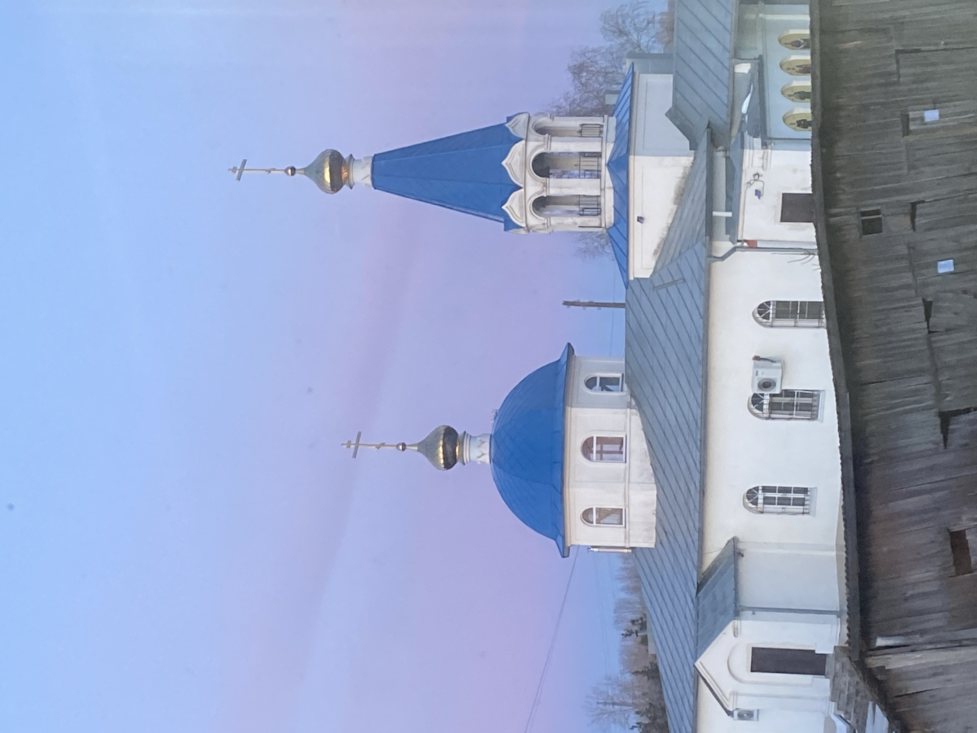 The image depicts a building with architectural elements characteristic of Russian Orthodox churches. It features a white building with blue rooftops and golden domes topped with crosses. The image is oriented vertically, with the church seemingly angled toward the top left corner. The sky is a pale blue with some violet hues, suggesting either dawn or dusk. A partial view of a wooden structure, possibly a wall or fence, is visible on the right side of the image. This architectural ensemble, with its distinctive domes and crosses, likely resides in Noviy, Russia, due to the question's reference to that city.