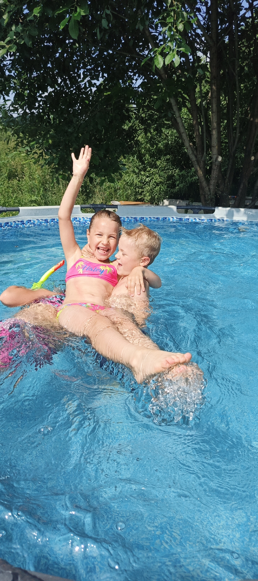 In this vibrant, sun-drenched image, two young children are joyfully playing in a bright blue swimming pool. The girl, wearing a pink bikini with a floral pattern, is seated and beaming, her arm raised in a wave. She appears to be holding onto the boy, who is nestled behind her, his arm wrapped around her shoulder and his head resting near hers. His expression suggests happiness and perhaps a bit of playful silliness. Both children are in the water, with splashes of water visible around their legs and feet, indicating active play.
The pool itself is surrounded by a white coping with a blue tiled edge. In the background, lush green trees and foliage create a natural, outdoor setting. The bright sunlight suggests it is a warm, clear day, likely midday or afternoon. The overall atmosphere is one of carefree childhood fun and summer enjoyment.
While no specific text is clearly legible on the children's attire, the scene conveys a strong sense of warmth, happiness, and summer recreation. The location context provided, Большой Букор, Russia, places this scene within a specific geographical area, suggesting a likely summer day there.