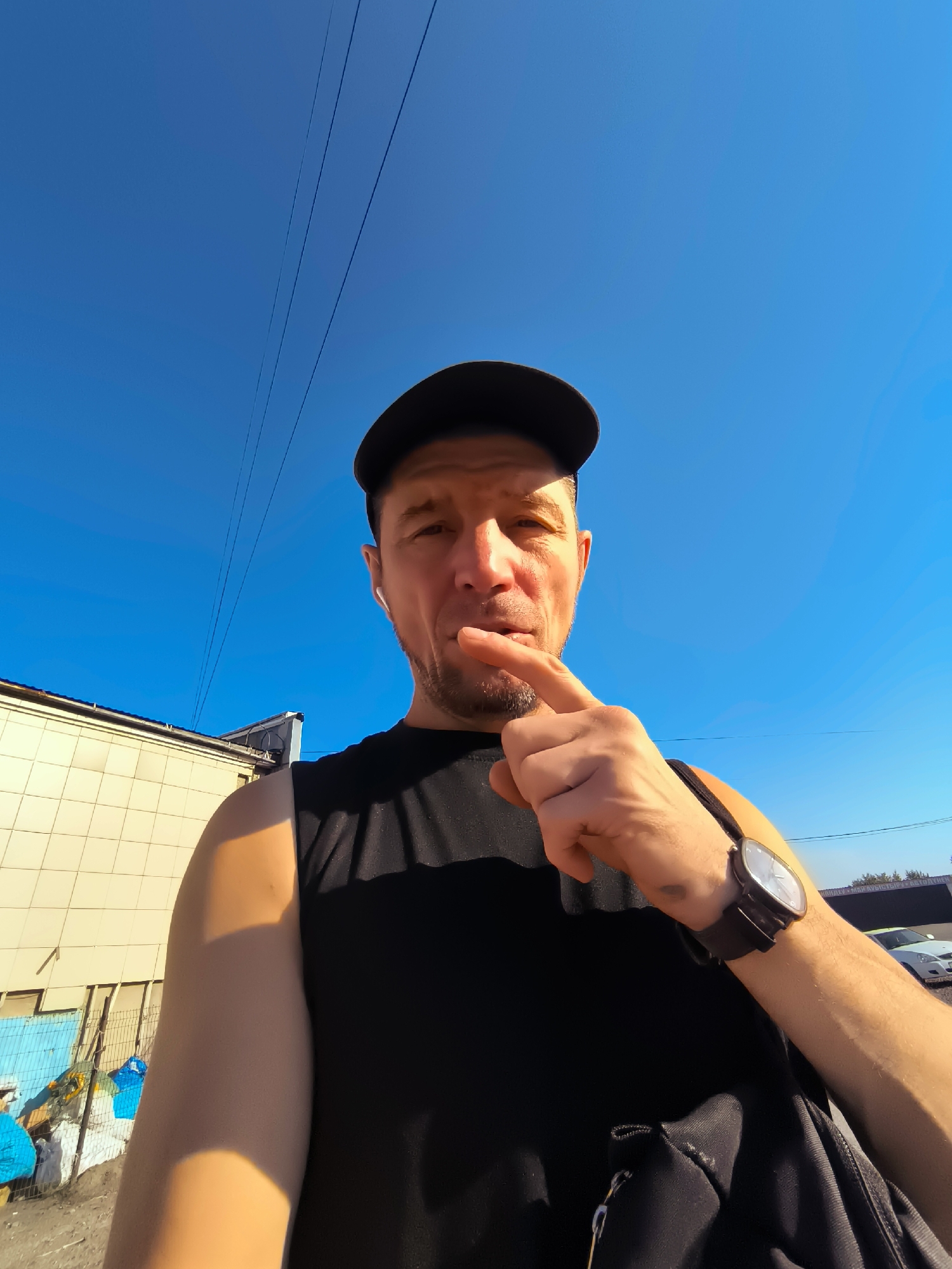 A low-angle, medium shot captures a man in a black baseball cap and a black sleeveless shirt, facing the camera outdoors on a bright, sunny day under a clear blue sky. He is looking directly at the viewer, with his right index finger touching his chin, in a contemplative pose. He is wearing a watch on his left wrist and appears to have a small, light-colored earbud in his right ear. Behind him, to the left, is a light-colored, tiled building. In the background, several utility wires stretch across the sky. The scene is set in an urban environment, with a chain-link fence and some debris visible in the lower left corner. The image quality is clear, with good lighting that casts shadows from the man and the building.