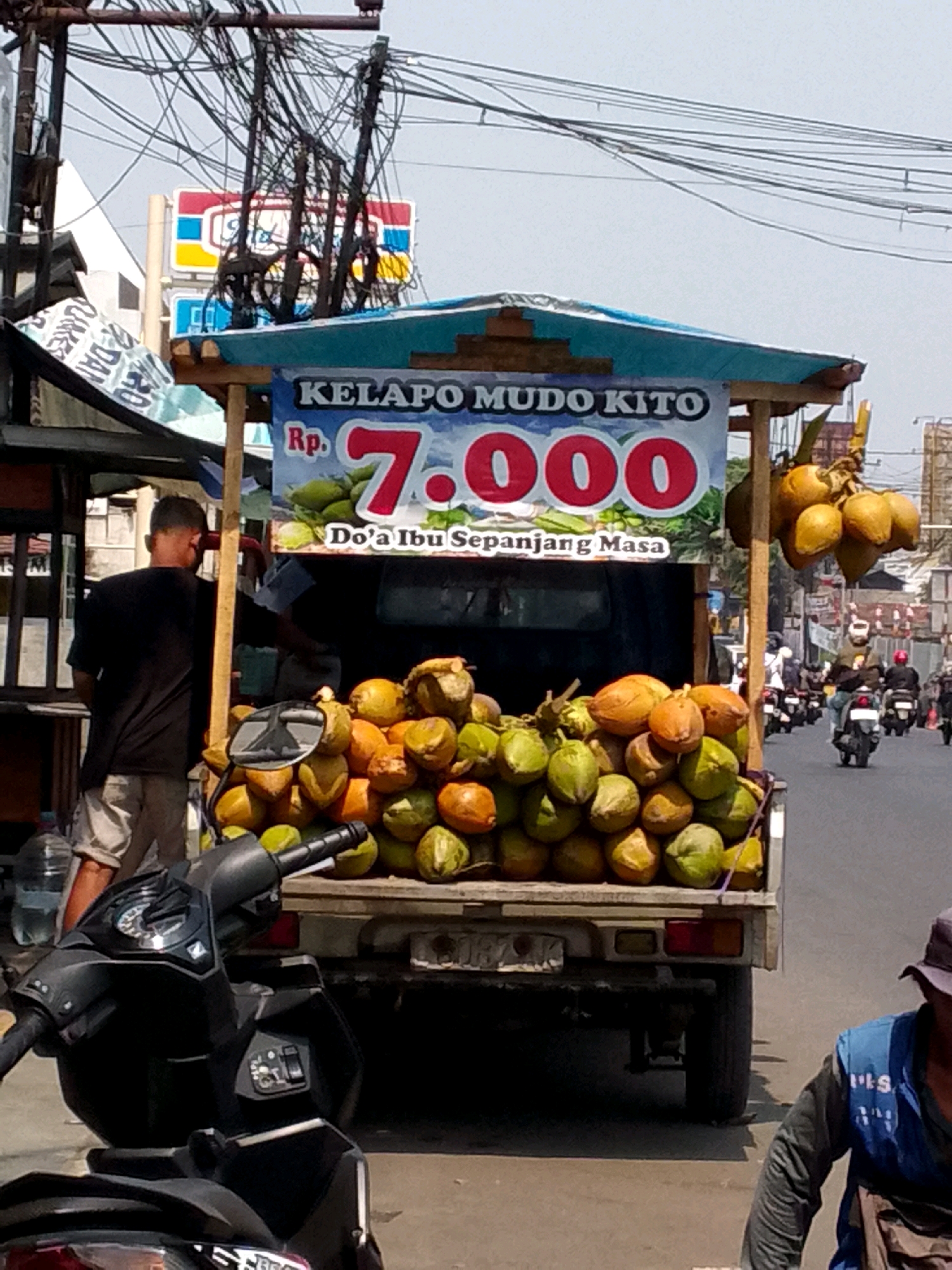A street vendor in Kertajaya, Indonesia, is selling young coconuts from a small truck. The truck is decorated with a banner advertising "KELAPO MUDO KITO" for Rp. 7.000. A large pile of coconuts, both green and brown, is visible on the back of the truck. A man in a black shirt and light-colored shorts stands behind the coconuts, appearing to be the vendor. In the foreground, a black motorcycle is parked. The street is busy with other motorcycles and a few pedestrians. The scene takes place on a sunny day, with overhead power lines and buildings visible in the background.