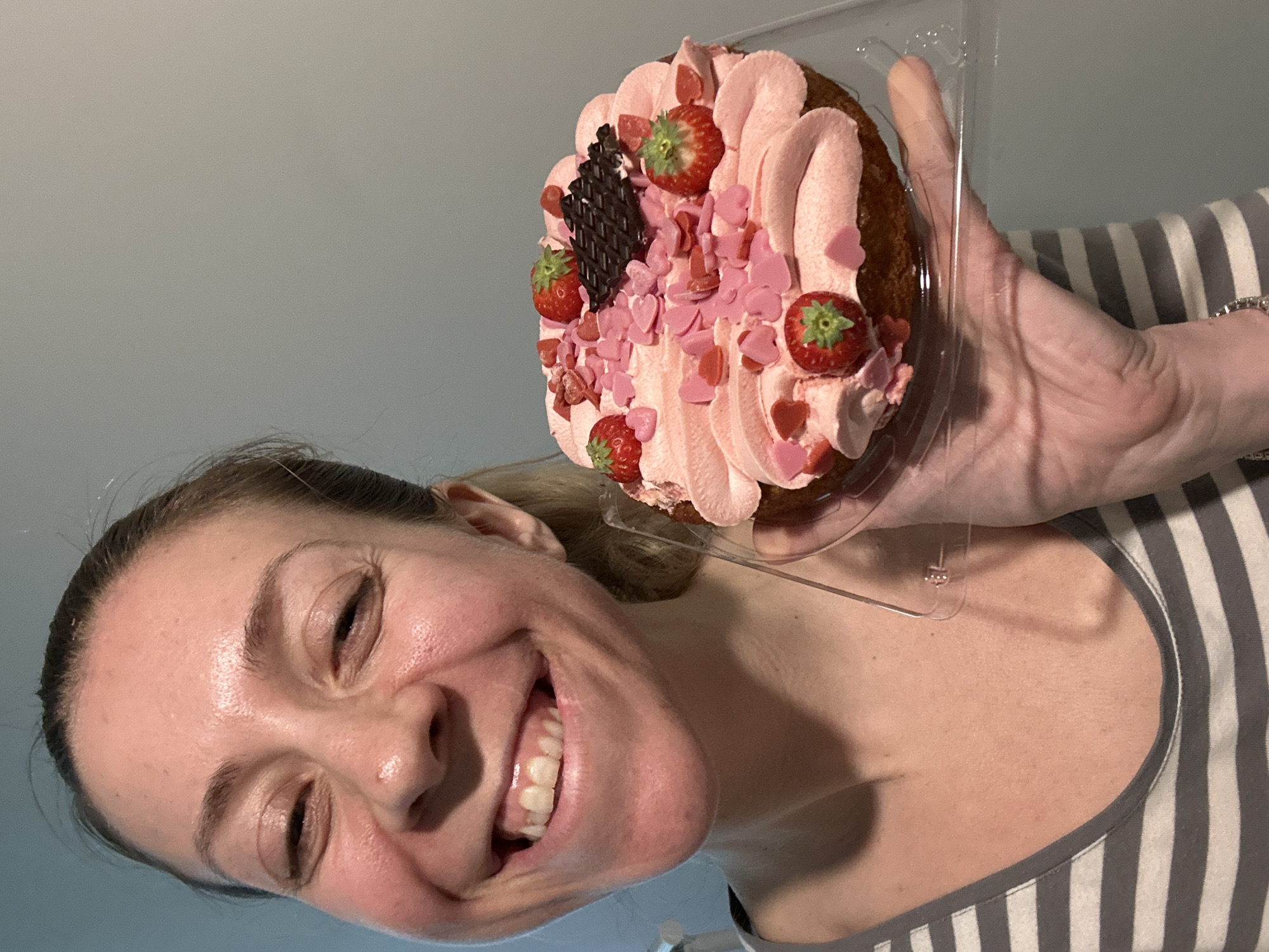 A woman with blonde hair pulled back smiles widely, holding a round cake in a clear plastic container. The cake is decorated with pink frosting, strawberries, and small pink heart-shaped sprinkles, with a small piece of dark chocolate placed on top. The woman is wearing a gray and white striped shirt. The background is a light gray wall. The image appears to be taken indoors. There is no indication of Amsterdam, Netherlands, in the image.