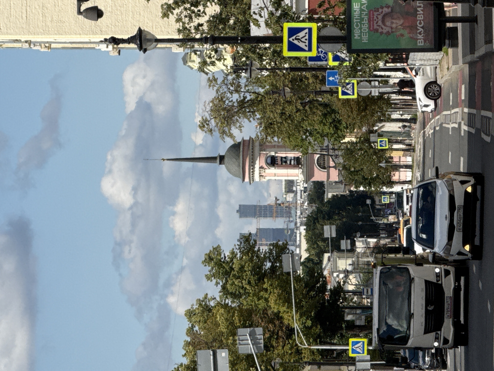 The image depicts a street scene in Moscow, Russia, likely in the daytime under a partly cloudy sky. A multi-lane road runs through the center of the frame, with several vehicles driving or stopped. On the left side of the road, a white truck is prominent, with a license plate partially visible. A white sedan is in the right lane, also with a visible license plate. Further down the road, other cars and a bus can be seen.
Traffic signs, including pedestrian crossing signs and directional signs, are visible along the roadside. Several trees with green foliage line the street. In the background, a pink and grey tower with a spire stands out, possibly part of a church or historical building. Beyond that, modern skyscrapers and construction cranes suggest urban development.
On the right side of the street, a tall billboard displays text and an image of a woman in traditional headwear. Streetlights and lampposts are also present. A few pedestrians are visible on the sidewalks, including one man in a black t-shirt. The overall impression is of a bustling urban environment with a mix of historical and modern architecture.