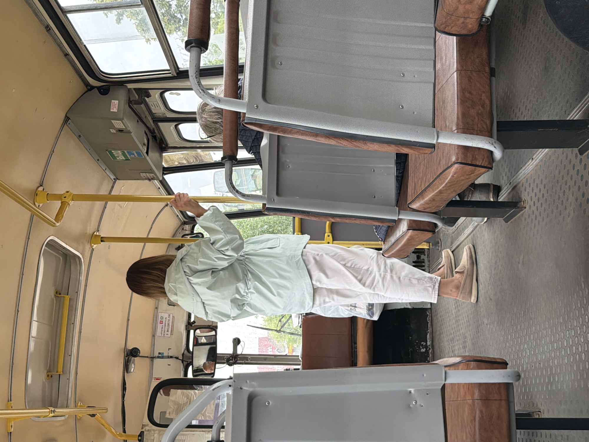 A person is standing inside a bus, holding onto a yellow handrail. They are wearing a light blue jacket and white pants. The interior of the bus is visible, with brown seats and yellow handrails. The bus appears to be in motion, as indicated by the view out the window. The lighting suggests it is daytime. The scene is set in Chaikovsky, Russia, as indicated by the location context. There is a sign on the wall with Cyrillic text, reading "АПТЕЧКА 100" and "У ВОДИТЕЛЯ", which translates to "First-aid kit 100" and "With the driver". Another sign with green text is also visible. The overall mood is neutral, with no strong emotional cues.