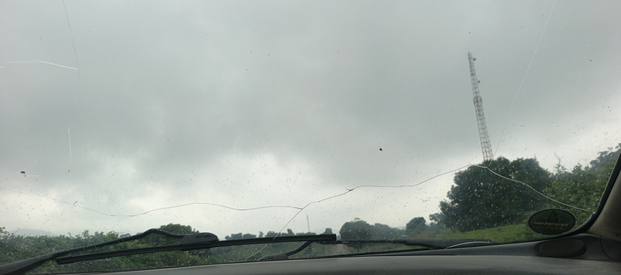 The image captures a view from inside a vehicle in Assob Hausa, Nigeria, looking out through a cracked and rain-speckled windshield on an overcast day. The car's dashboard and windshield wipers are visible in the foreground.
A prominent, large crack traverses the windshield horizontally across the middle and lower sections, with several finer cracks also apparent, particularly in the upper left. Numerous small water droplets or specks of dirt are scattered across the glass.
Beyond the windshield, the sky is a uniform, dull grey, indicative of heavy cloud cover. On the right side of the frame, a tall, red and white telecommunications tower rises above the tree line. The midground consists of a dense, green expanse of trees and foliage, suggesting a rural or natural landscape.
No people are visible. An oval-shaped green sticker, with text that is too indistinct to read, is attached to the dashboard area on the right, near the windshield frame. The scene conveys a moment of observation from a car, under gloomy weather conditions, with the damaged windshield being a central element of the visual narrative.