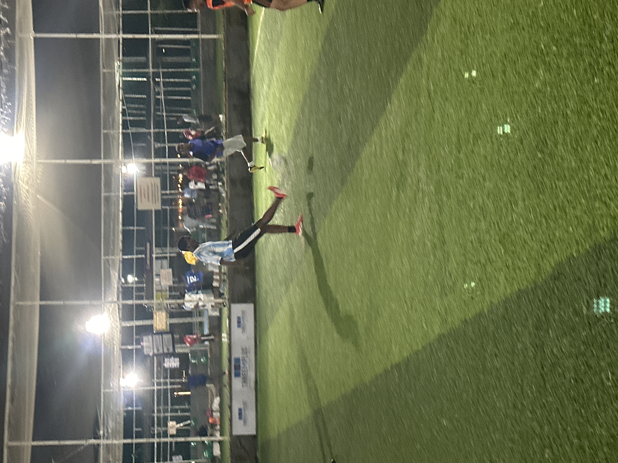 The image depicts an outdoor artificial turf soccer field at night, illuminated by overhead floodlights. The field has a dark green surface with lighter green stripes. In the foreground, a person wearing a light blue and white striped jersey and black shorts with white stripes on the side is in the process of kicking a soccer ball. They are wearing pink cleats. The person's shadow is cast on the ground.
In the background, several other individuals are visible, some appearing to be players and others spectators. They are standing near a white netted enclosure that surrounds the field. Signs are mounted on a structure behind the enclosure. The overall environment suggests a sports facility in an urban setting. Based on the presence of the "Abuja International Soccer Academy" banner, this scene is located in Abuja, Nigeria.