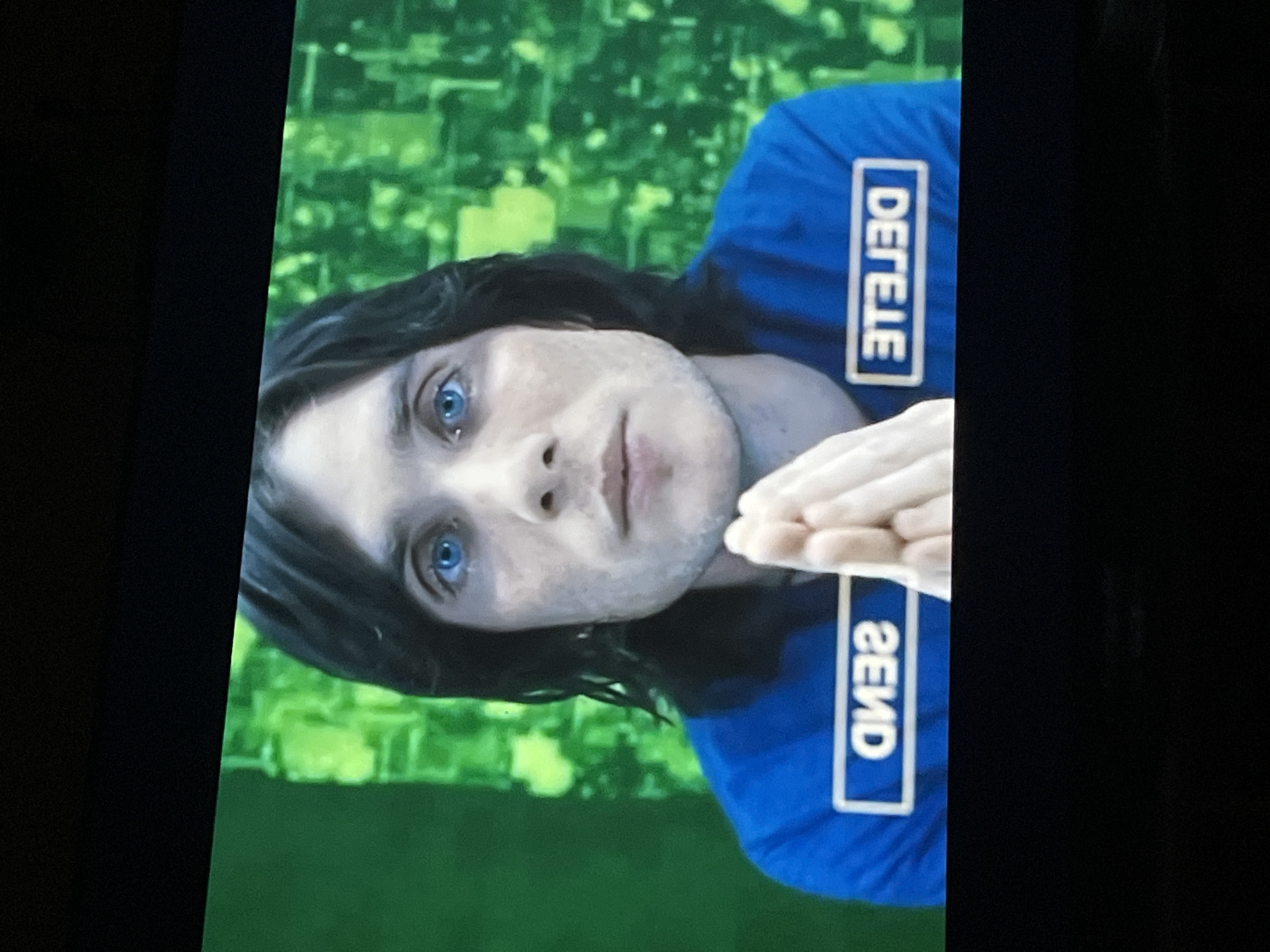 The image shows a person with light skin, dark hair, and blue eyes. They are wearing a blue shirt with the words "DELETE" and "SEND" vertically printed on it. The background is a blurry green, resembling a digital or surveillance screen. The image appears to be taken indoors, possibly from a television or monitor. The location of Baan Mae Nam, Thailand, is not depicted in the image.