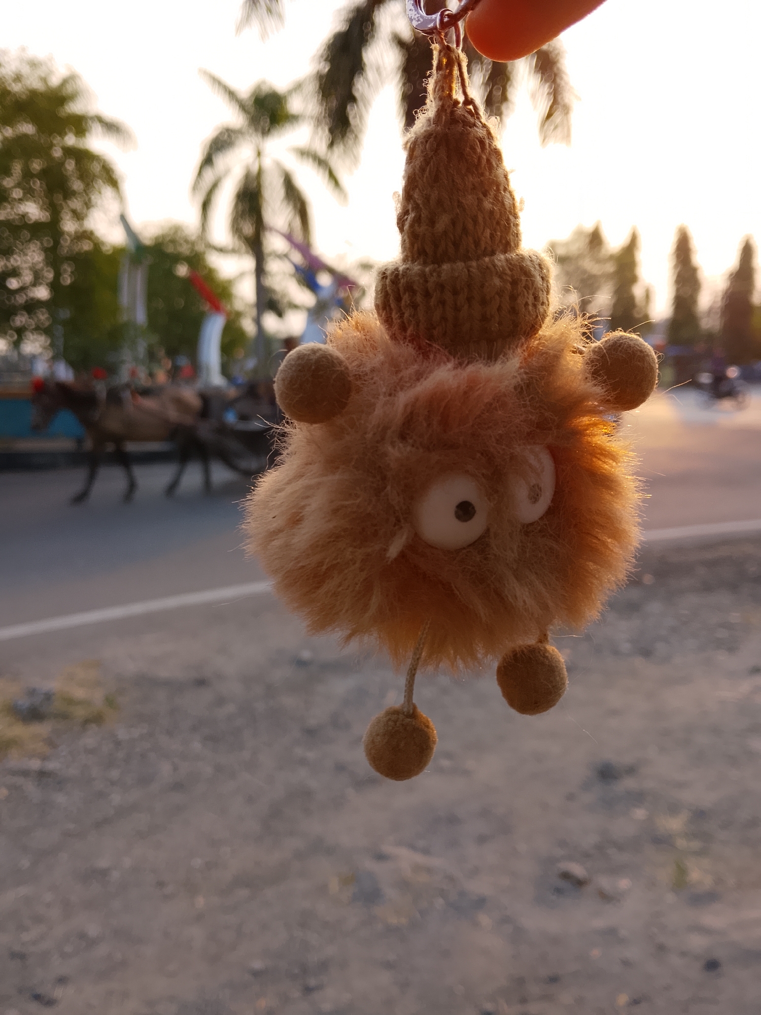 A fuzzy, brown, cartoonish creature keychain hangs in the foreground against an outdoor, sunlit backdrop. The creature has large, googly eyes, a knitted brown cap, and two small pom-poms dangling from its body. The background, slightly out of focus, shows a road with a white line, a blurred silhouette of trees, and what appears to be a horse-drawn cart in motion. The overall lighting suggests it is either sunrise or sunset. The location is specified as Balun, Indonesia.