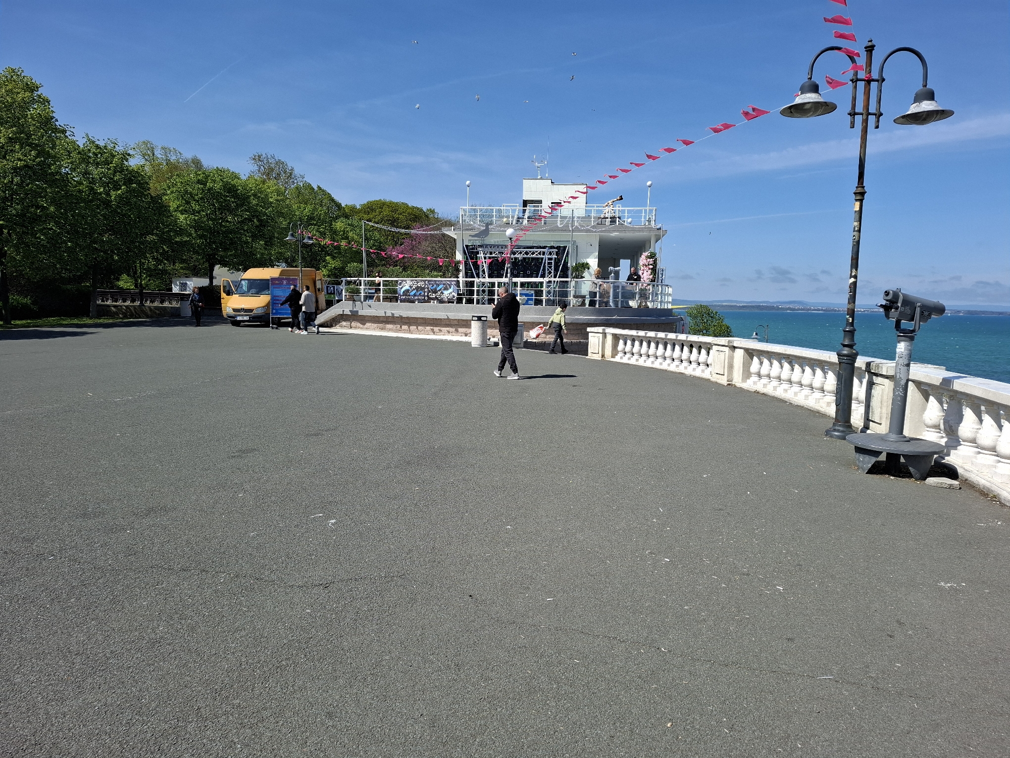 The image shows a sunny day on a paved area overlooking the sea. A building with a white railing is set up as a stage or event venue, adorned with red pennant flags. There are people on and around the building. A yellow van is parked nearby. A person in dark clothing stands in the middle of the paved area, possibly taking a picture. A child in a light-colored jacket walks beside them. To the right is a lamppost with three lamps and a coin-operated telescope. The paved area leads to a white stone balustrade, beyond which is the sea. Green trees line the left side of the image. This is likely the area known as the Sea Garden in Burgas, Bulgaria, given the coastal setting and type of architecture visible.