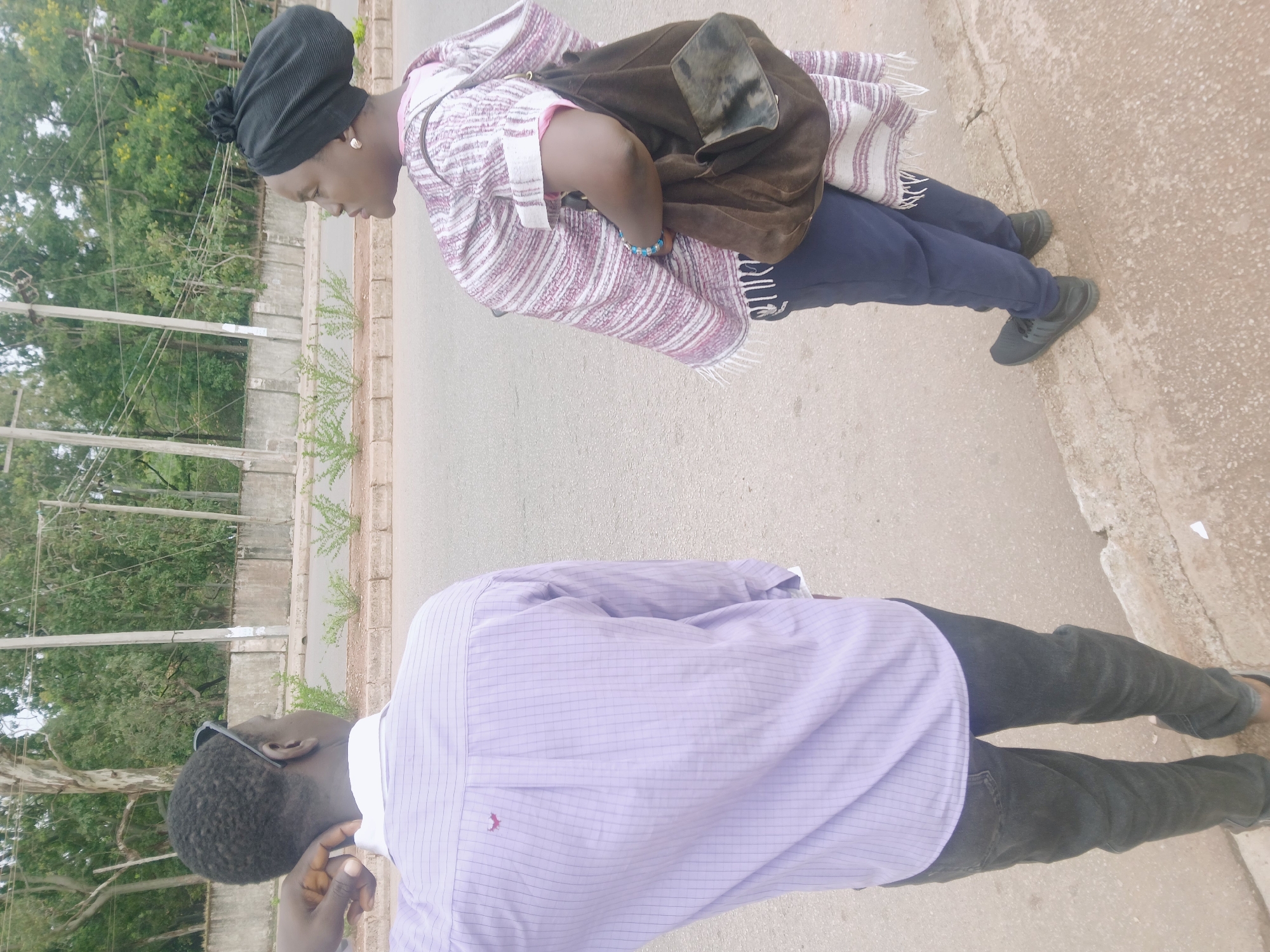 A partially obscured view shows two individuals standing on a paved road in what appears to be the city of Bukuru, Nigeria. The individual closest to the top of the frame is wearing a striped shawl or garment, dark pants, dark shoes, and a black head covering, with a brown bag slung over their shoulder. The other individual is wearing a light purple, long-sleeved button-down shirt, dark pants, and dark shoes. Vegetation and a low wall are visible on the left side of the frame. Electrical poles are visible as well. The road surface appears worn and weathered.