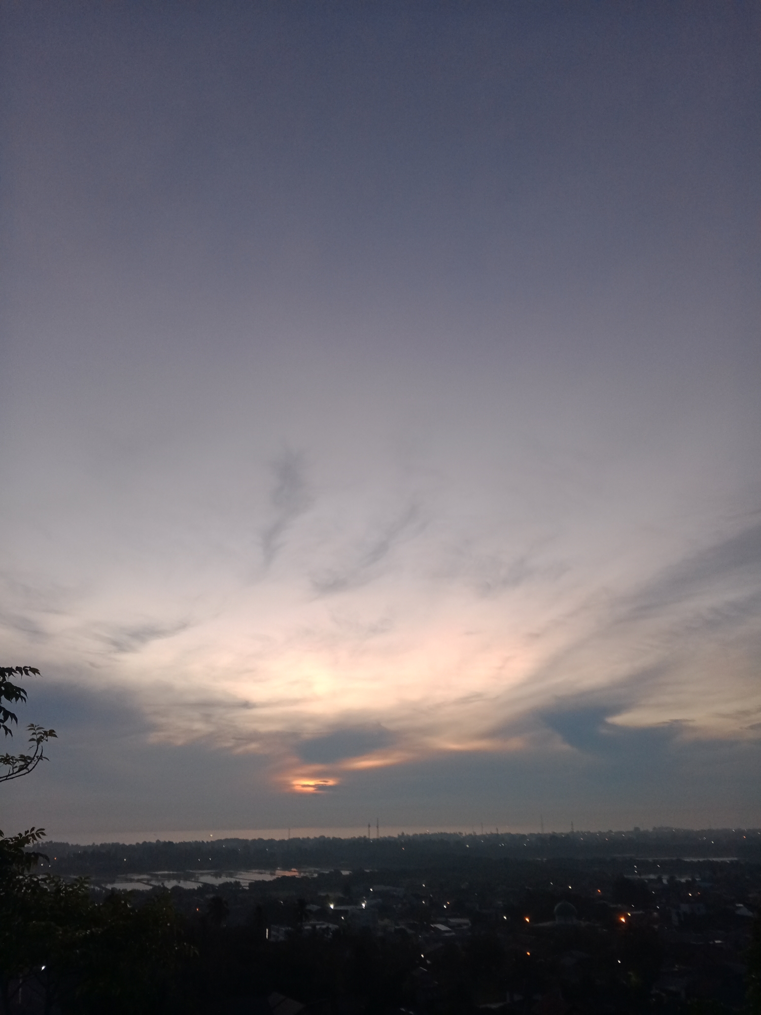 This image captures a serene dawn sky over the city of Lhokseumawe, Indonesia. The sky is a canvas of soft grays and muted purples, transitioning into a warm, ethereal glow near the horizon where the sun is beginning to rise. Wispy clouds stretch across the sky, some catching the nascent light and appearing as streaks of pink and orange. Below the expansive sky, the city of Lhokseumawe stretches out, a silhouette of buildings, trees, and a distant body of water. The cityscape is dotted with faint lights, suggesting the early hours before the city fully awakens. In the foreground, dark foliage frames the view, adding a sense of depth and natural beauty to the urban landscape. The overall atmosphere is one of tranquility and the gentle awakening of a new day.