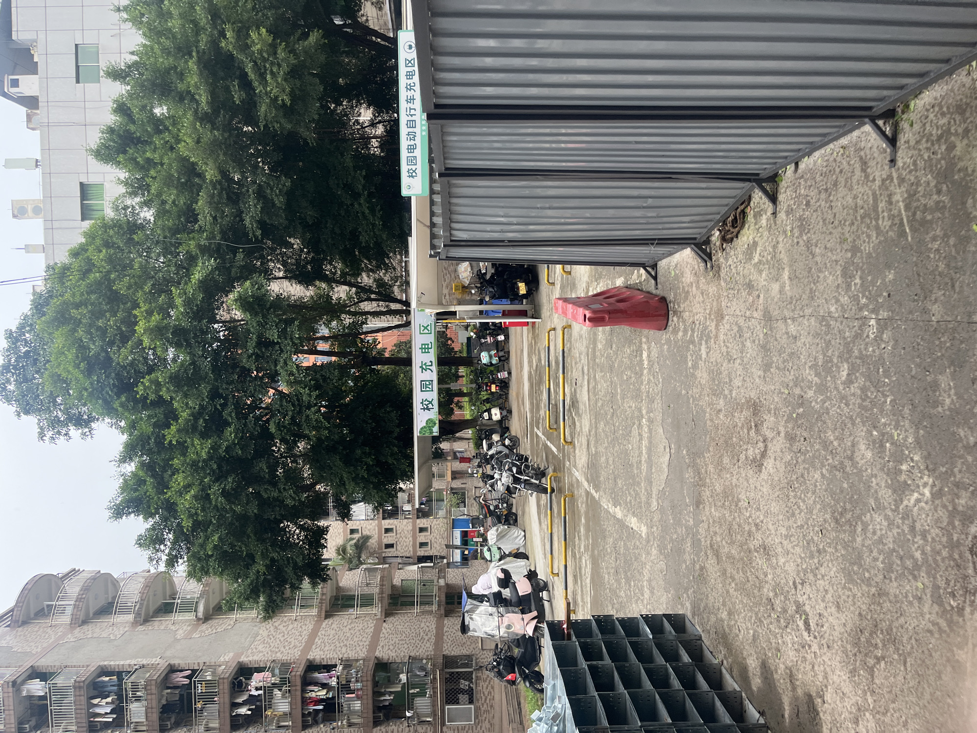 The image depicts a parking area for electric scooters and motorcycles in Yuzhong District, China, likely on a cloudy day. In the background, a multi-story apartment building with balconies adorned with hanging laundry stands prominently. Adjacent to it, a dense cluster of trees forms a green canopy. A covered charging station for electric vehicles is visible, marked with Chinese signage that translates to "Campus Electric Vehicle Charging Area." The foreground shows a paved lot with several scooters and motorcycles parked. A red plastic barrier and a grey corrugated metal fence are also present. To the left, a dark grey metal shelving unit is positioned. The ground surface is wet and appears to be concrete, with some scattered leaves. There are no people directly visible in the image.