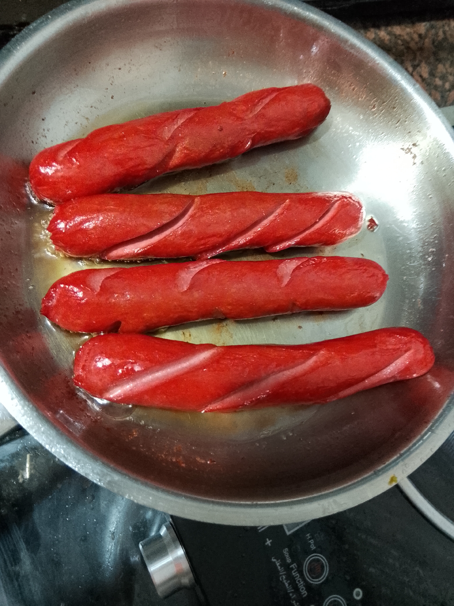 The image shows four red sausages with diagonal cuts, placed in a circular metal pan with oil. The sausages are arranged horizontally, one above the other. The pan is on a black stovetop, which appears to be an induction cooker, as indicated by the control panel visible at the bottom of the image with text in English and Arabic, including "Soup Function" and "H. Pot". The environment is a kitchen. There is no visible indication of the location being in Davao City, Philippines.