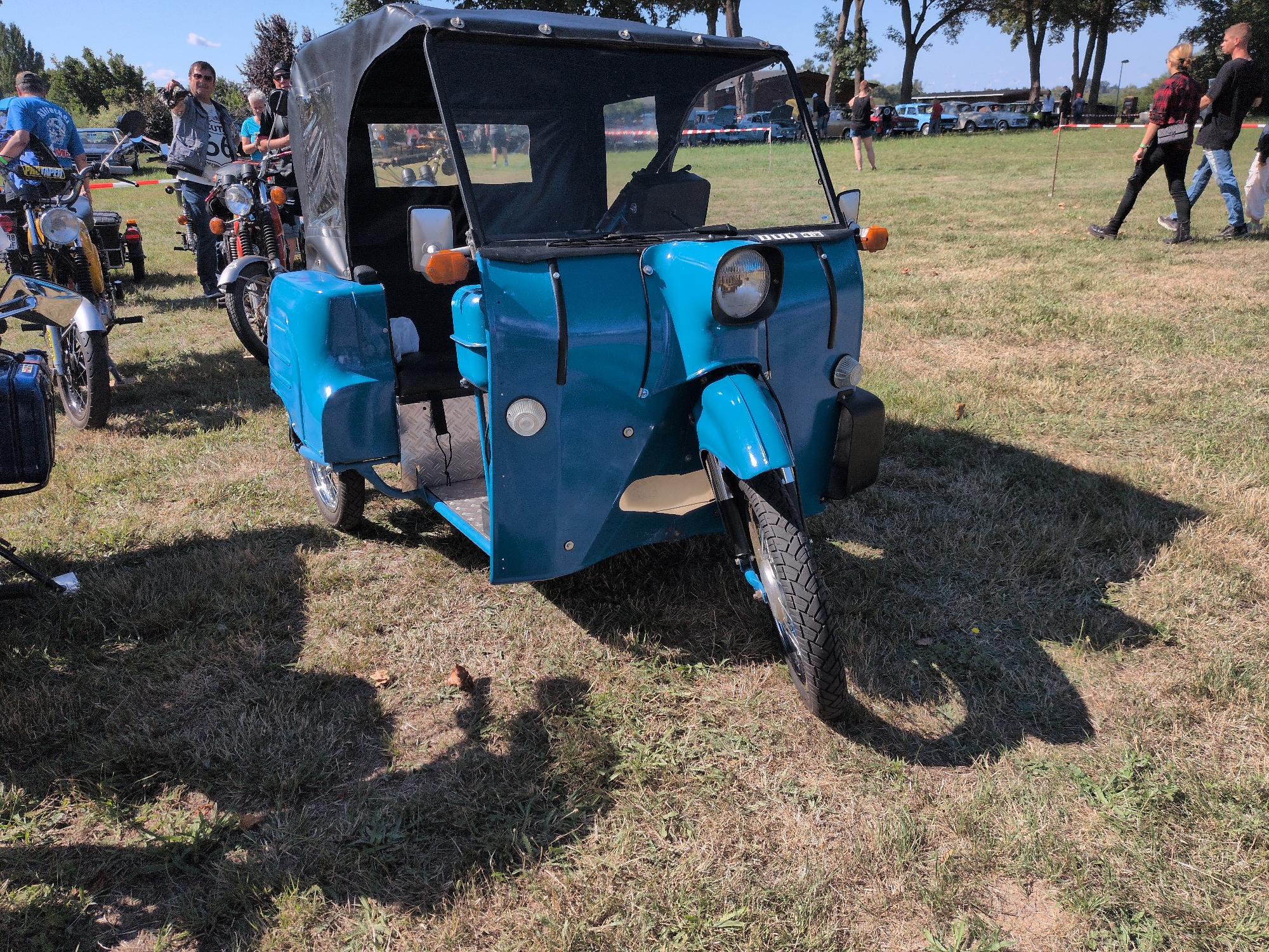 This is a medium shot, eye-level, outdoor photograph of a blue three-wheeled vehicle parked on a grassy field. The vehicle is a vintage-style auto rickshaw with a black fabric canopy. The front of the vehicle features a single large headlight, turn signals, and a blue fender. The interior of the vehicle is partially visible, showing a black seat and metal footplates. The ground is covered in dry grass and shadows are cast by the vehicle and people in the background.
In the background, several people are visible. To the left, a man in a blue shirt and jeans stands next to a motorcycle. Further back, a man in a grey vest and jeans points to the right. Other people and several older model cars are scattered throughout the background. The sky is clear and blue. The location is identified as Hohenferchesar, Germany.