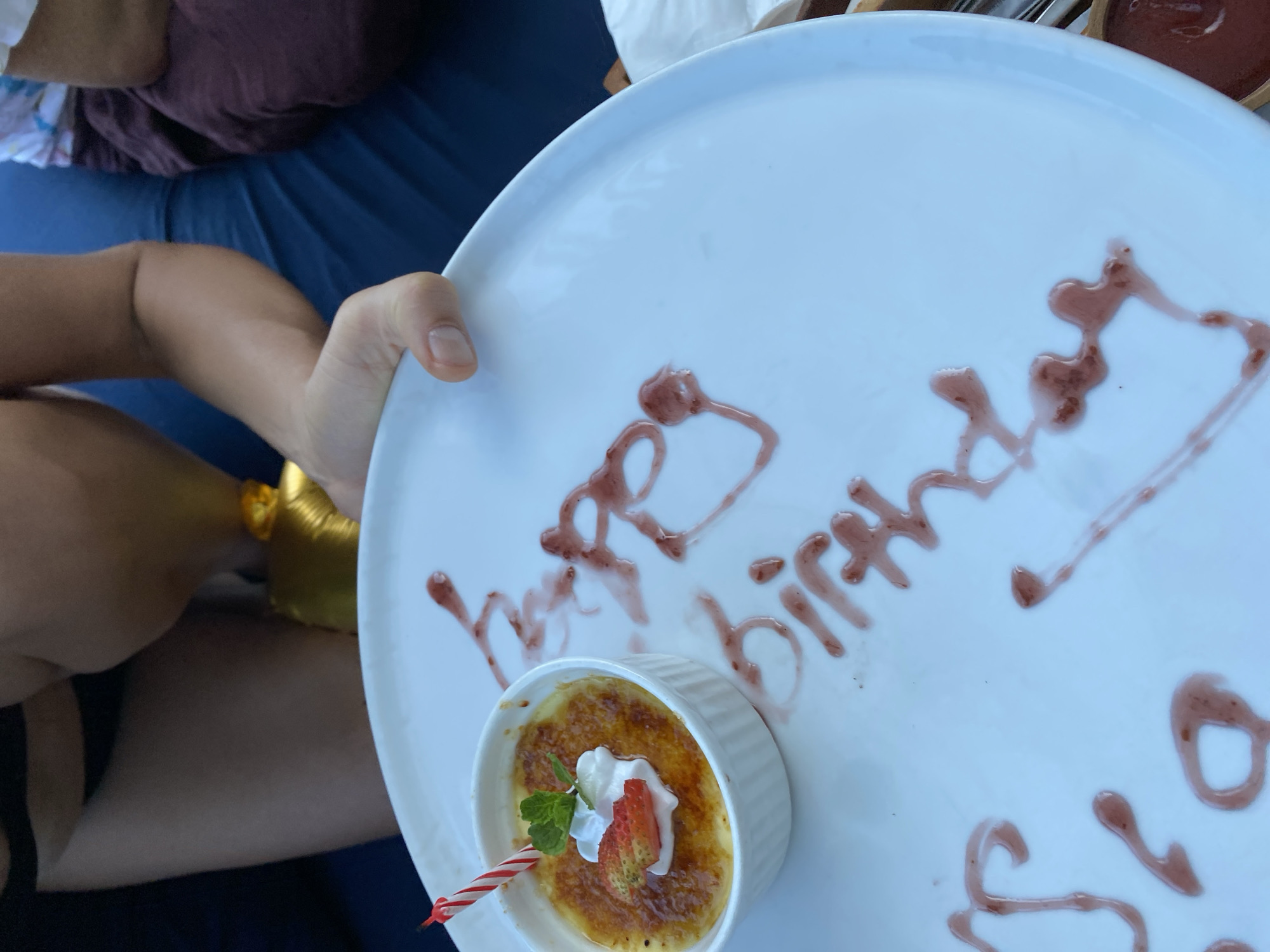 The image displays a white plate with a creme brulee dessert topped with whipped cream, a strawberry slice, and a lit birthday candle. The dessert is in a white ramekin. The plate is decorated with red sauce, forming the words "Happy Birthday". A person's hand is visible holding the plate. The background shows a person wearing dark clothing and sitting on a blue surface. The scene is outdoors, likely in Kuta Selatan, Indonesia, given the context provided. The lighting suggests it is daytime.