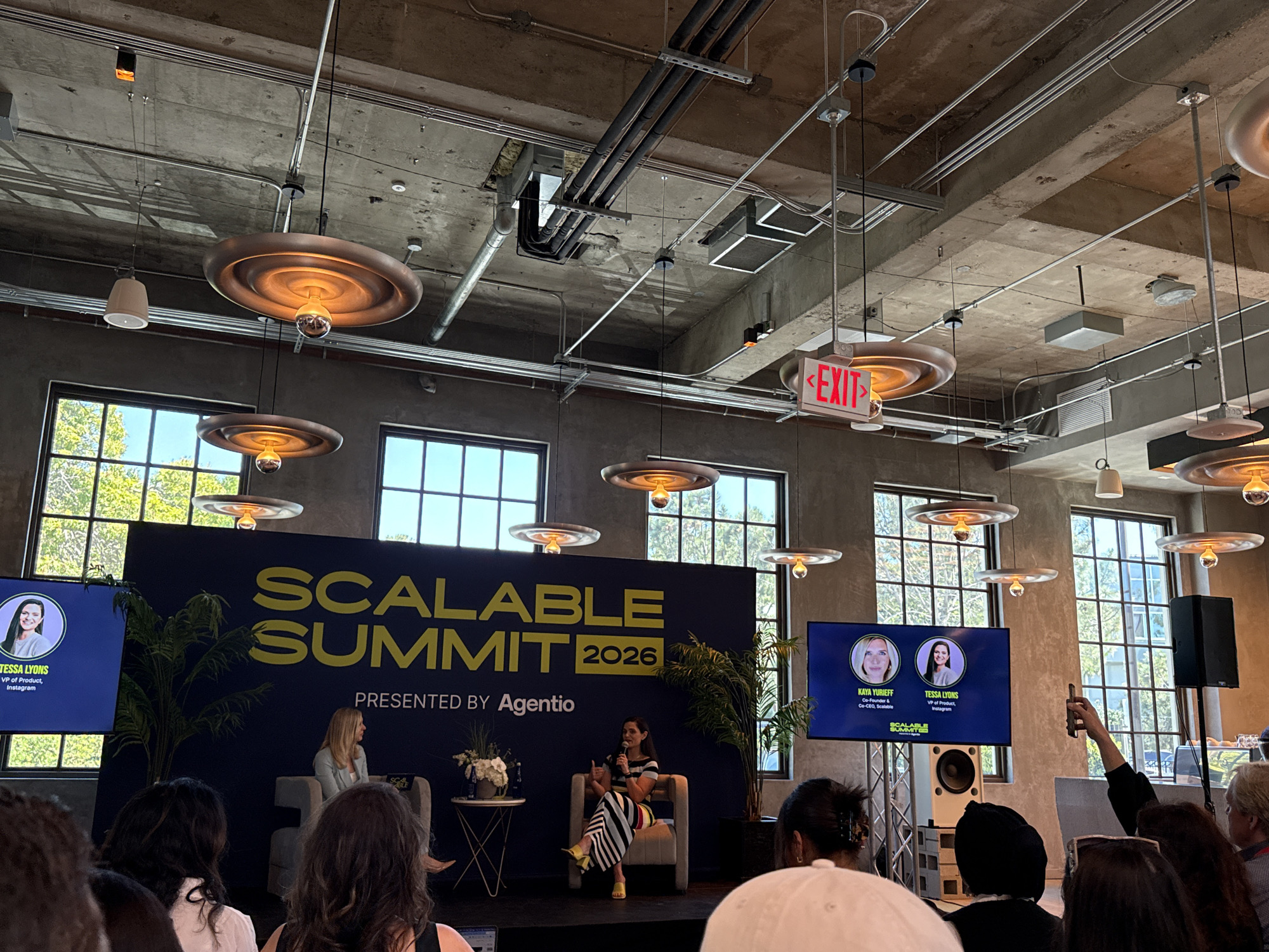 The image captures a scene from the Scalable Summit 2026, presented by Agentio, which is taking place in a city, most likely Los Angeles, United States of America. The event features a panel discussion with Tessa Lyons, VP of Product at Instagram, and Kaya Yurieff, Co-Founder & Co-CEO of Scalable, as indicated by the screens on either side of the stage.
The stage is set against a backdrop banner displaying the event's name and sponsor. Two individuals, presumably Lyons and Yurieff, are seated in chairs on stage, engaged in conversation with one holding a microphone. A small table with flowers sits between them.
An audience is seated in front of the stage, with attendees visible in the foreground. One audience member is holding up a smartphone, possibly to take a photograph or video of the event.
The environment features an industrial-style ceiling with exposed pipes and ducts, as well as pendant lights. Large windows behind the stage provide natural light and a view of the outside. There's an "EXIT" sign hanging from the ceiling above the stage. Speakers are placed on either side of the backdrop.