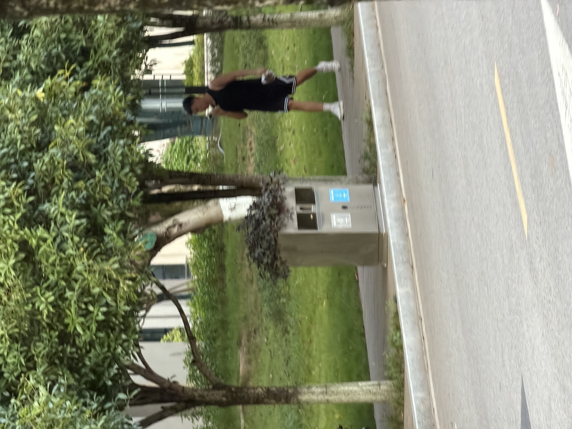 The image captures a young man in a black sleeveless shirt and black shorts with white stripes walking on a paved sidewalk next to a grassy area. He is holding a white object in his right hand and appears to be speaking on a phone held to his left ear. To his left, there is a grey trash receptacle with what looks like a plant on top. Trees with dense green foliage are visible in the background, as well as a building. The scene appears to be an outdoor urban setting, possibly a park or a street with landscaping. The lighting suggests it is daytime. The image's metadata does not specify a location in Jiangjin, China, but the general environment is consistent with many urban areas in China.