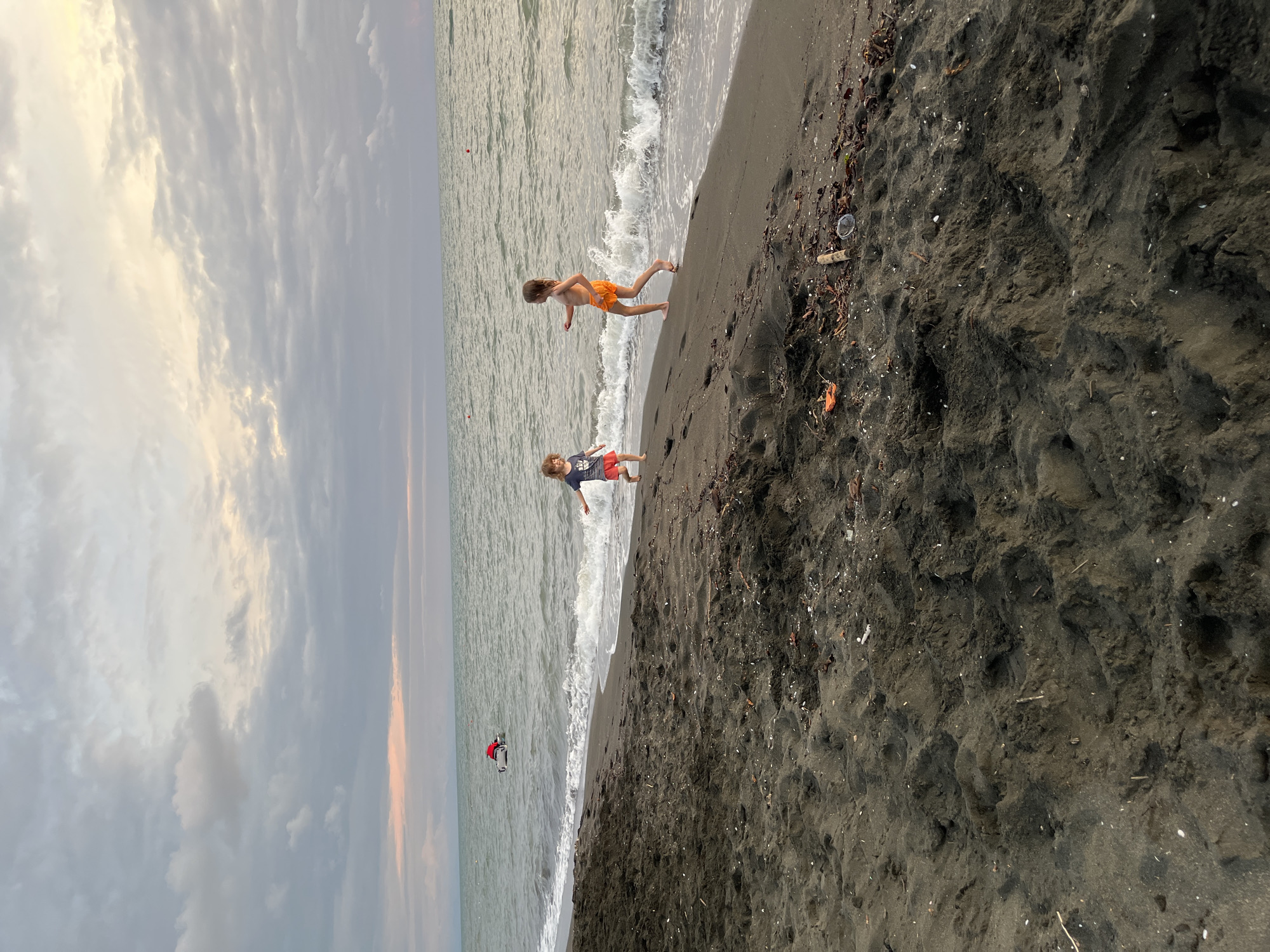 Two children are playing on a dark sand beach near the Black Sea in Ureki, Georgia. The child on the left, wearing a striped t-shirt and red shorts, is running towards the water. The child on the right, wearing orange shorts, is facing away from the camera and running along the shore. The sea is calm with small waves breaking on the beach. In the distance, a person is on a small boat with a red top. The sky is overcast with some pastel colors visible near the horizon. The beach is covered in dark sand with some scattered debris.