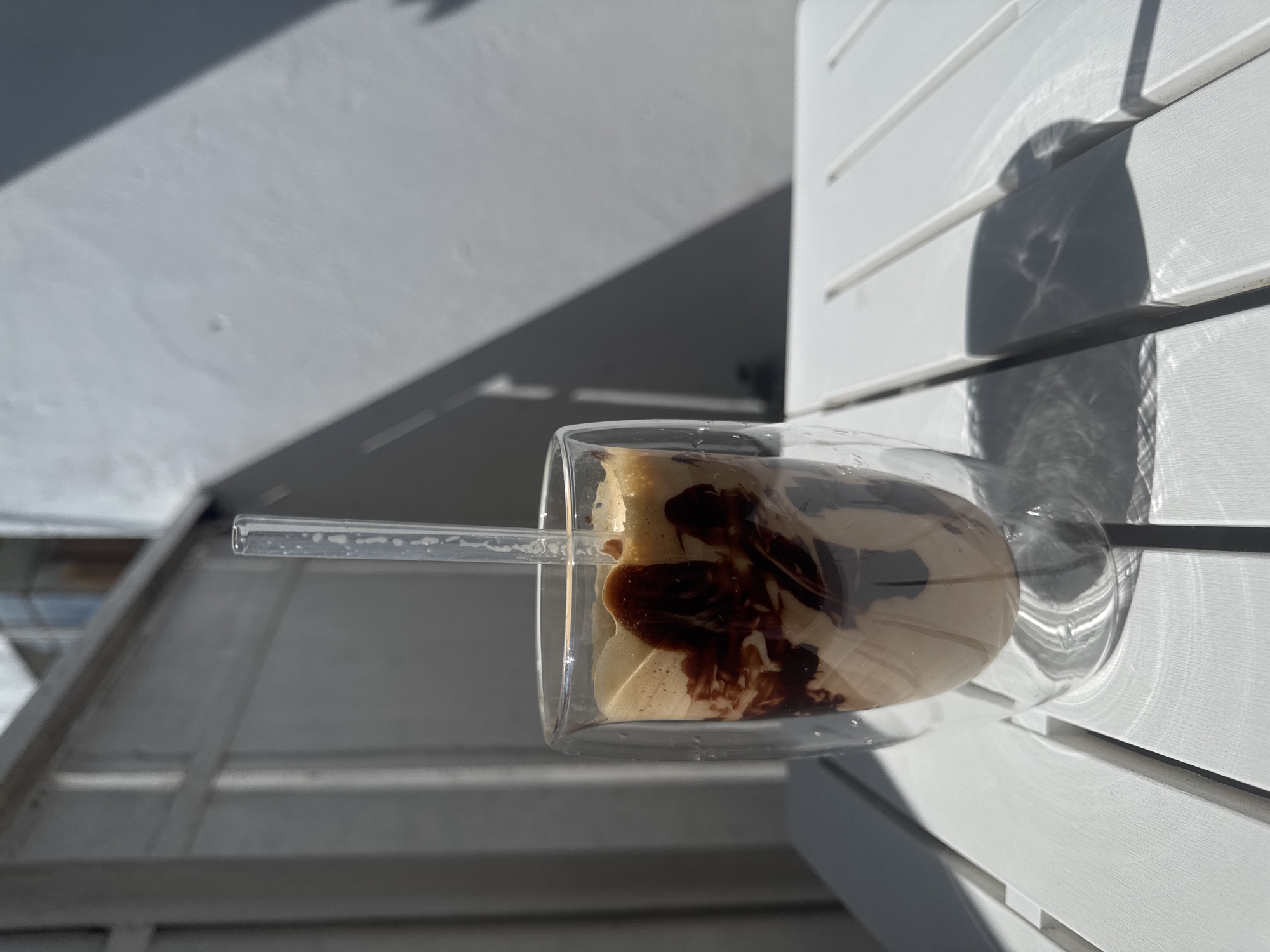 A clear glass with a drink inside, likely coffee, sits on a white slatted surface. The drink is layered with a light brown foamy substance and a darker brown syrup, possibly chocolate, drizzled on top. A clear straw extends from the drink. The glass is oriented horizontally in the frame. The background shows a light-colored, textured surface with a diagonal shadow cast across it, suggesting it is outdoors and the sun is shining. To the right of the glass, a white slatted structure is visible, also with shadows. The overall lighting and shadows indicate it is a sunny day. The location is Varna, Bulgaria. There is no visible text.