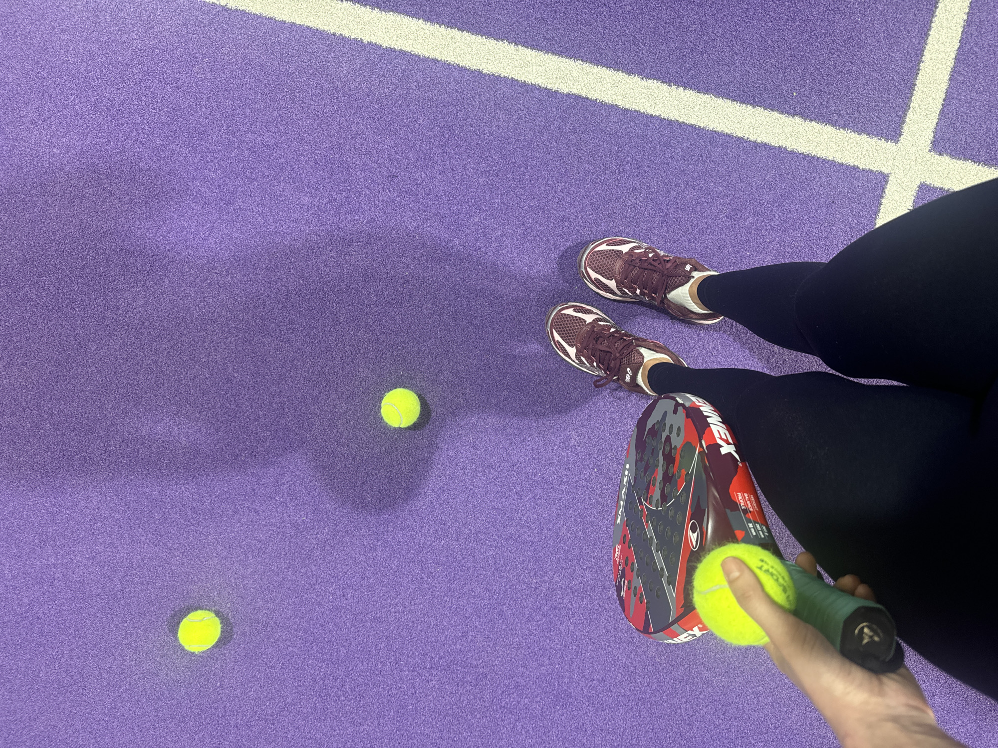 The image shows a person standing on a purple padel court with white boundary lines. The person is wearing dark leggings and burgundy sneakers. There are two tennis balls on the court surface, and the person is holding a padel racket and another tennis ball. The city is not visible and therefore cannot be identified.