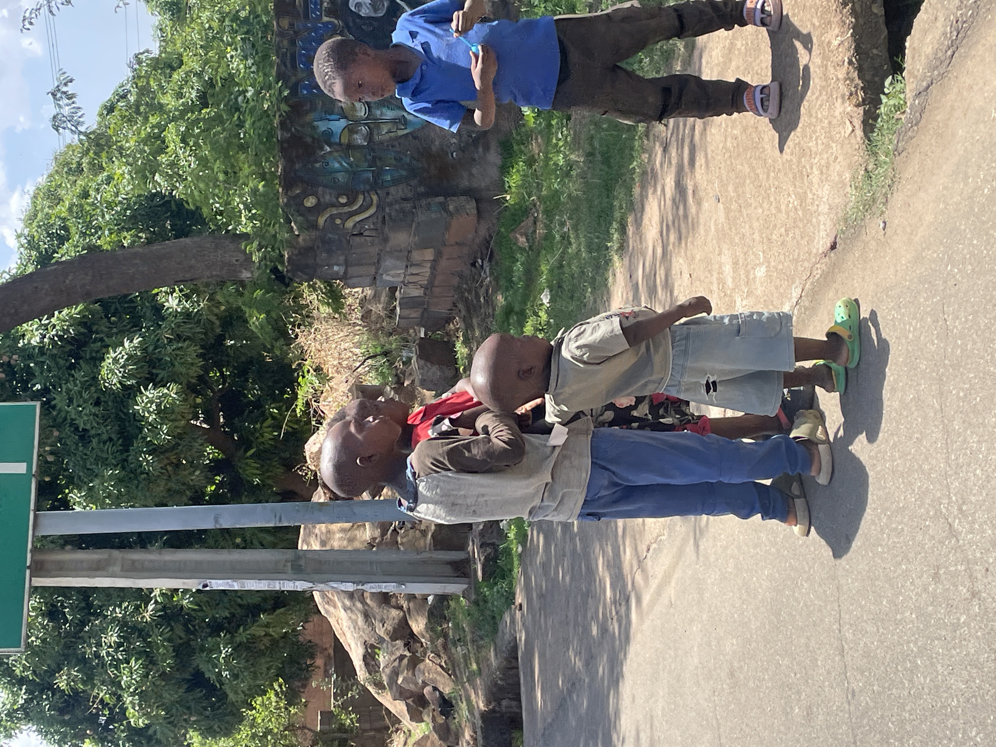 The image depicts a street scene in Jos, Nigeria, featuring four young boys. Two boys are positioned on the right side of the image, one wearing a blue shirt and brown pants, blowing bubbles. The other two boys are standing closer to the camera; one wearing a khaki shirt and blue shorts and the other wearing blue pants and a light-colored shirt. The ground is paved and there are trees and greenery visible in the background, as well as a partially visible sign on the left side of the image. A concrete wall with colorful graffiti is located behind the boys blowing bubbles. The overall setting suggests a neighborhood or residential area within the city.