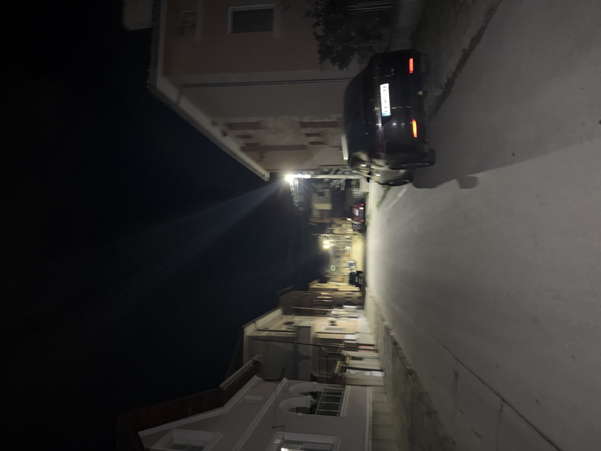The image depicts a street in Panagyurishte, Bulgaria, at night. A dark-colored car is parked on the right side of the road, facing away from the viewer. The street stretches into the distance, flanked by buildings on both sides. Streetlights illuminate the road, and a cluster of buildings can be seen further down the street. The sky on the left is dark.