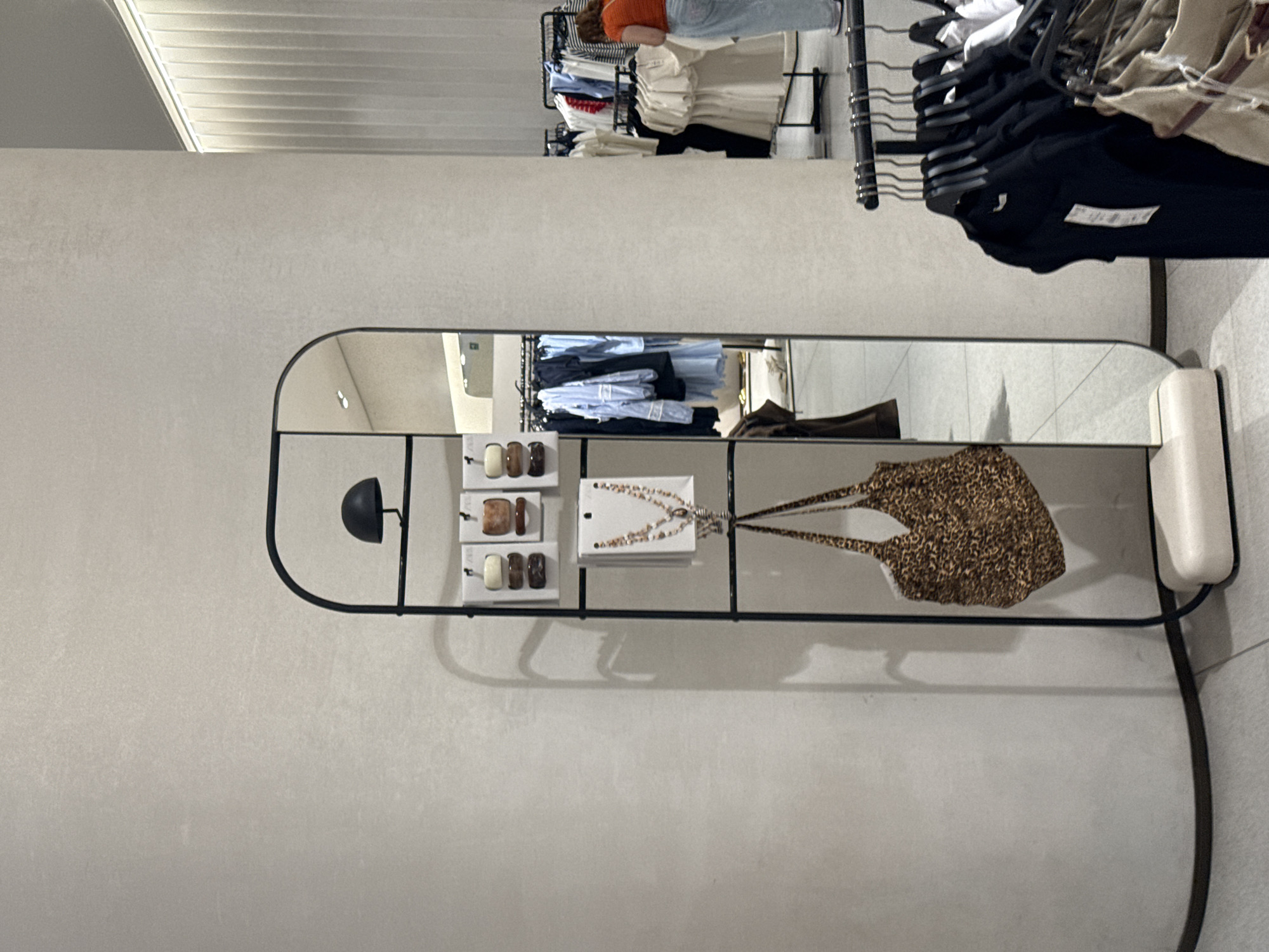 The image shows a display rack with various items for sale, including jewelry, a leopard print bag, and other accessories. The reflection in the mirror behind the rack reveals clothing on hangers, and what appears to be a person. The scene appears to be inside a retail store. There is no information to suggest that this is located in the city of Puebla, Mexico.