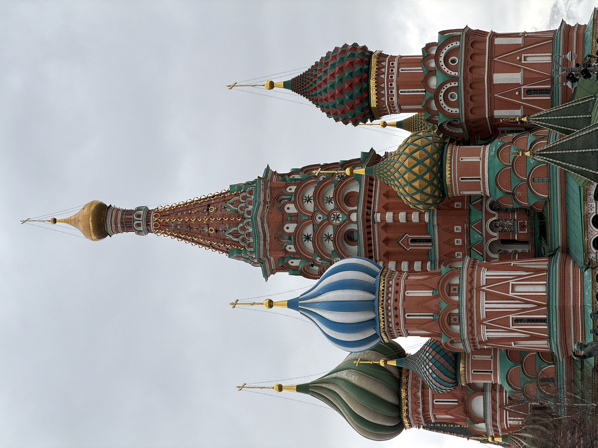 The image shows St. Basil's Cathedral in Moscow, Russia. The cathedral features multiple uniquely decorated domes with distinctive patterns and colors. The building is red brick with white and green accents. The sky is overcast, suggesting a cloudy day. No people are distinctly visible in the image, although there are some individuals visible at the top of the structure in the background.