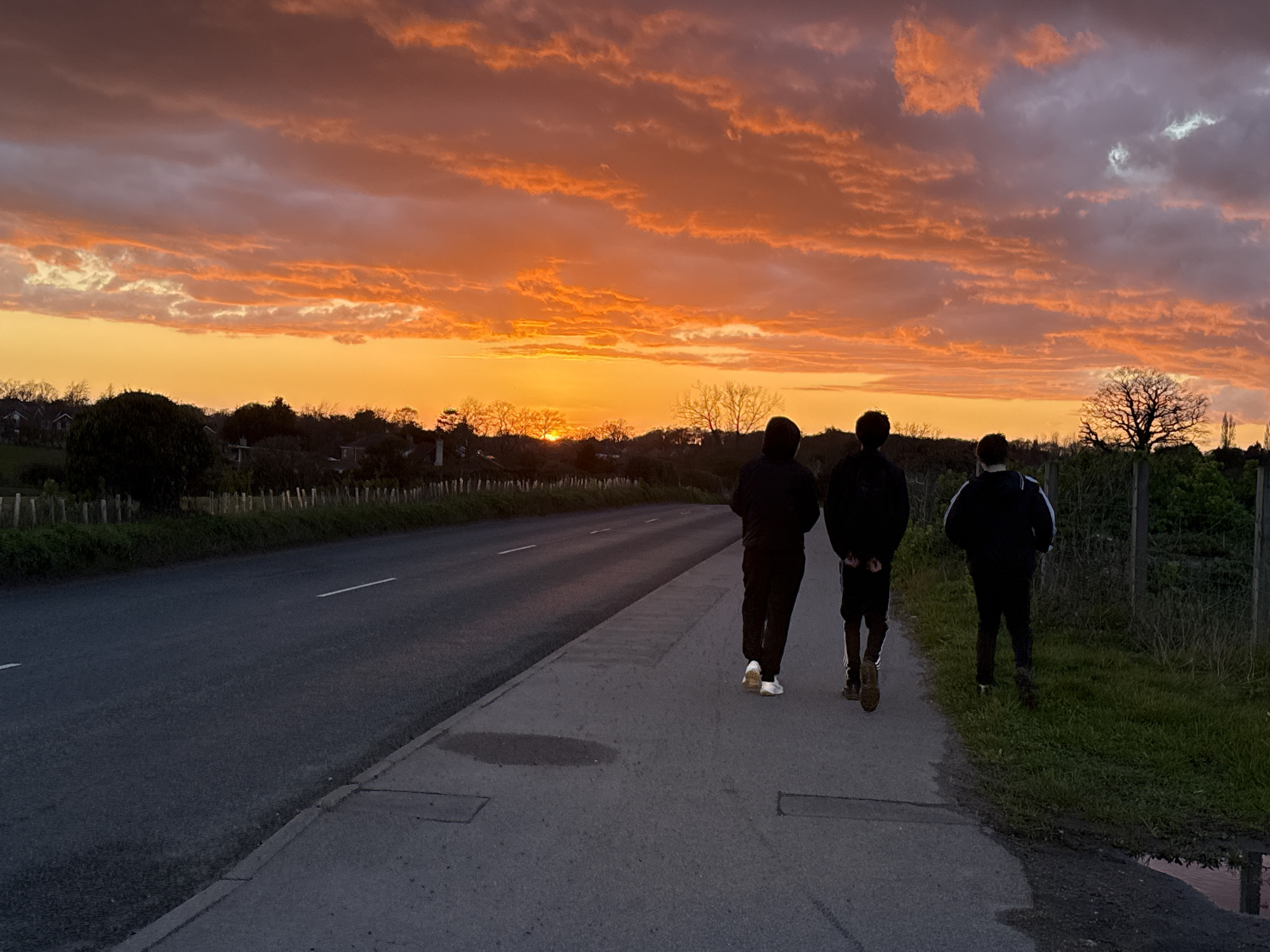 Three people are walking away from the viewer along a paved sidewalk adjacent to a road. The road has a single white line marking the center. Beyond the road, there is a fence and some trees. The sky is a vivid orange color, indicating a sunset or sunrise. The location is unspecified, and cannot be definitively determined as Great Yarmouth, United Kingdom from the image.