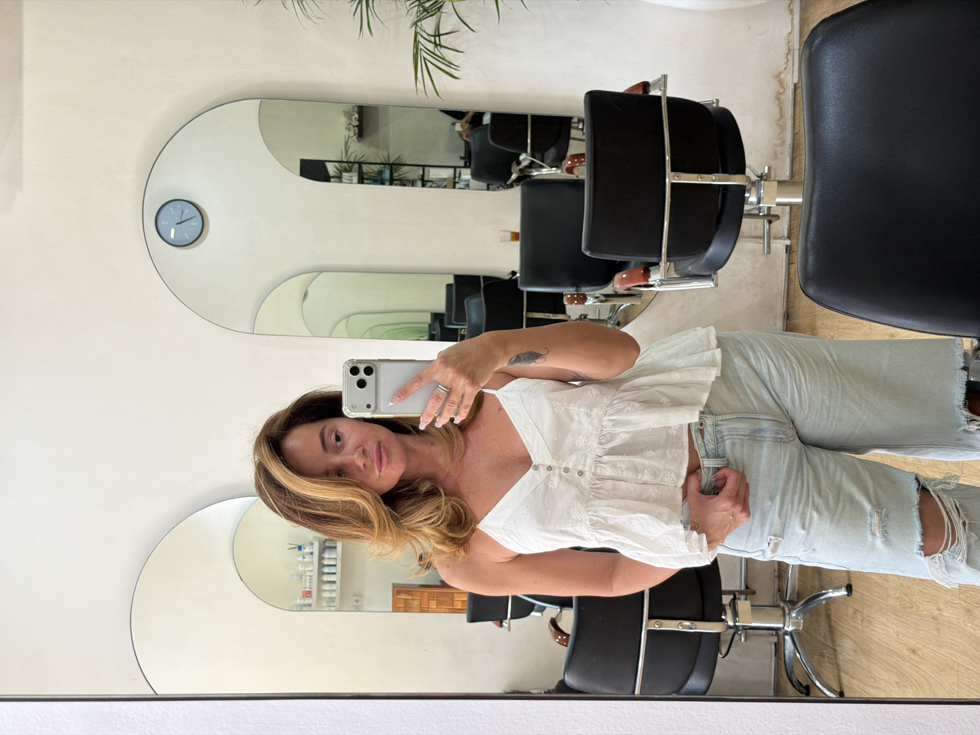 A medium-shot shows a woman taking a selfie in a hair salon in North Kuta, Indonesia. The woman is wearing a white, embroidered top and light-wash ripped jeans. She is holding a white smartphone in her right hand, angled slightly towards the mirror. Her hair is a mix of blonde and brown. The background features a series of curved mirrors, reflecting the salon's interior with black salon chairs and hair products on shelves. A wall clock is visible on the wall next to a mirror. The salon has a minimalist and modern design.