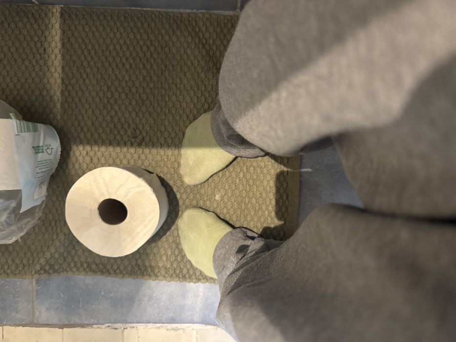 A downward-facing first-person view captures the lower extremities of an individual standing on an olive-green, textured mat. The individual wears grey sweatpants and light green socks. Centered directly in front of their feet on the mat is a single roll of white toilet paper. In the upper right quadrant of the image, a partially visible clear plastic bag containing a multi-pack of paper product, likely toilet paper, displays labeling that includes "FSC C104523" and a green recycling-like symbol. The mat rests on a floor tiled with light blue or grey-blue square tiles. A sharp shadow line runs diagonally across the mat and the individual's socks, indicating a distinct light source. The scene is located in Paris, France.