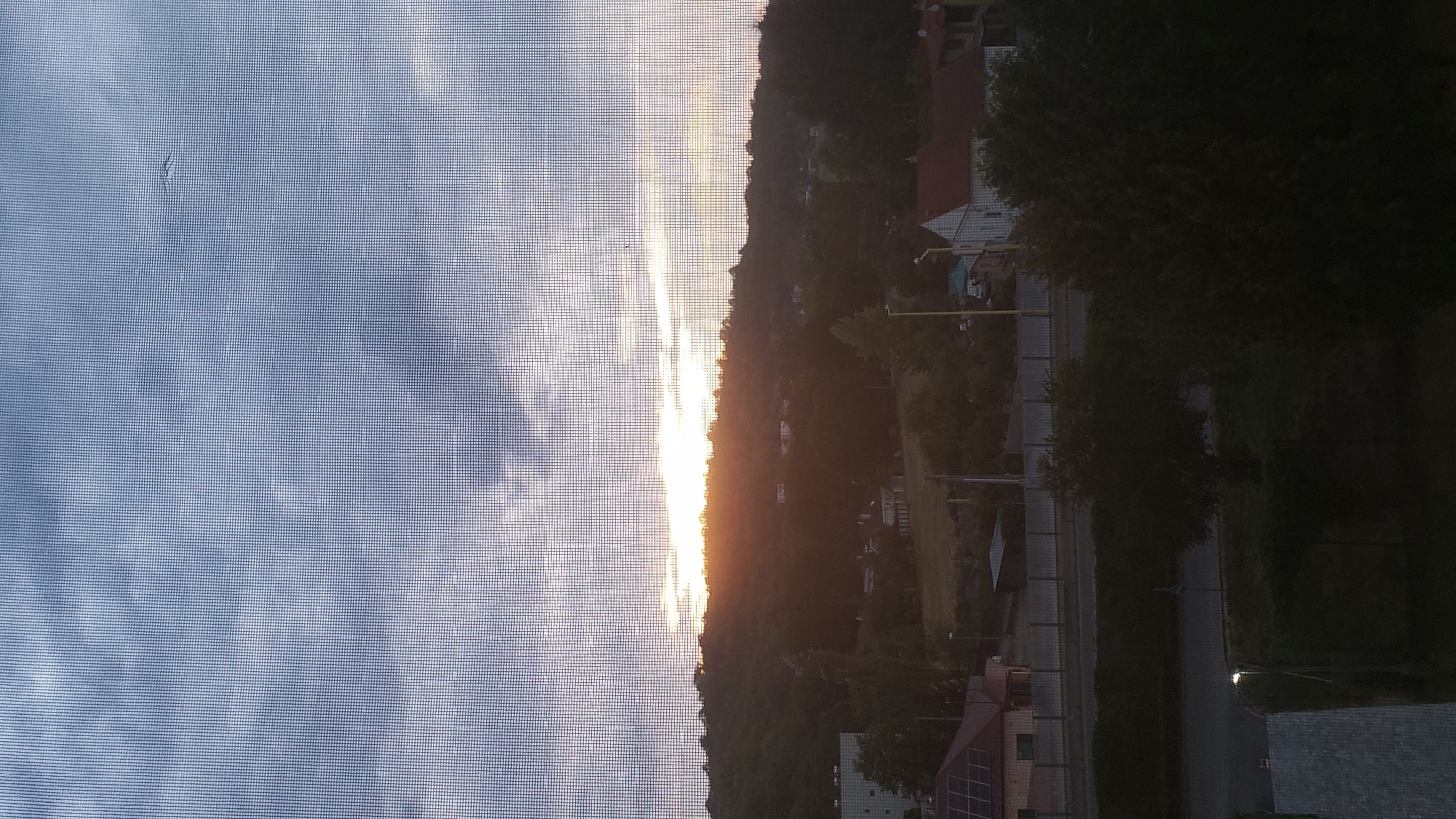 The image depicts a view through a window screen of a sunset over a residential area in Prešov, Slovakia. The sky is filled with clouds, illuminated by the setting sun which casts a warm glow over the landscape. In the foreground, the window screen creates a grid pattern, obscuring some of the details.
Below the sky, a forested hill dominates the middle ground, with several houses and buildings scattered among the trees. The houses appear to be of varying sizes and architectural styles, some with red tiled roofs. A road runs along the base of the hill, and a few vehicles are visible. The foreground is a dark silhouette of trees and buildings, with a street lamp visible on the left side.
The overall impression is one of a peaceful evening in a suburban or semi-rural setting. The scene is calm and serene, with the natural beauty of the sunset contrasting with the man-made structures of the town.