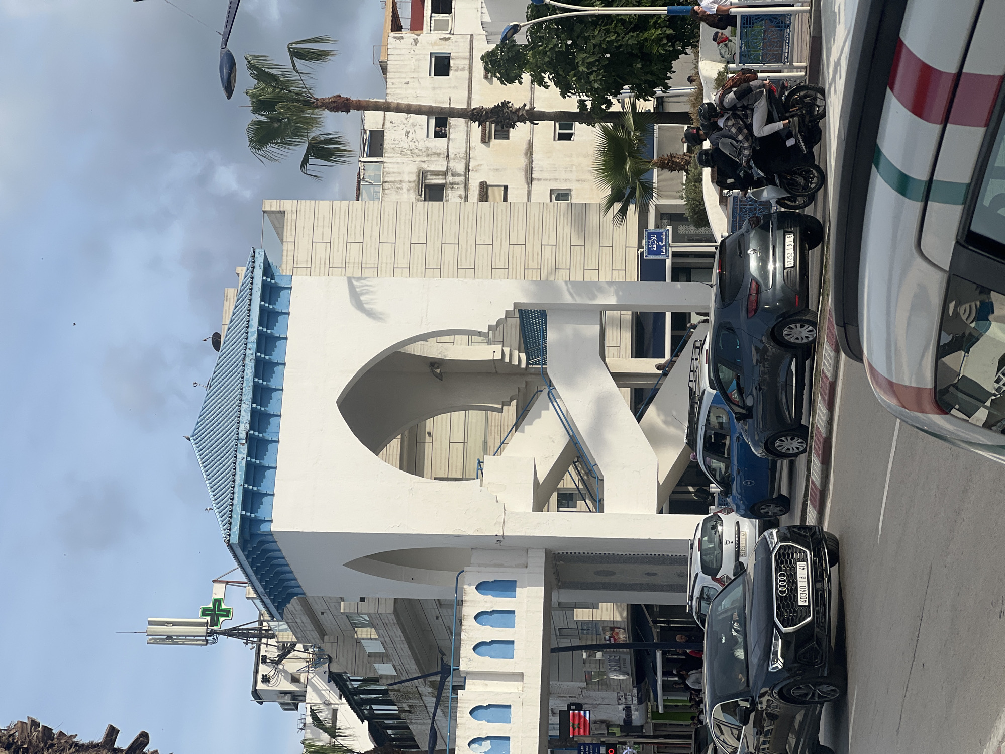 The image depicts a street scene in the city of M'Diq, Morocco. The primary architectural structure in view is a white building with a blue-tiled roof and a large circular opening. Adjacent to it is a taller, off-white building. Palm trees are visible in the background. The street is lined with parked cars including a black Audi. A motorcycle with multiple riders is present. Street signs are also visible. A pharmacy sign is also visible. The sky is partially cloudy.