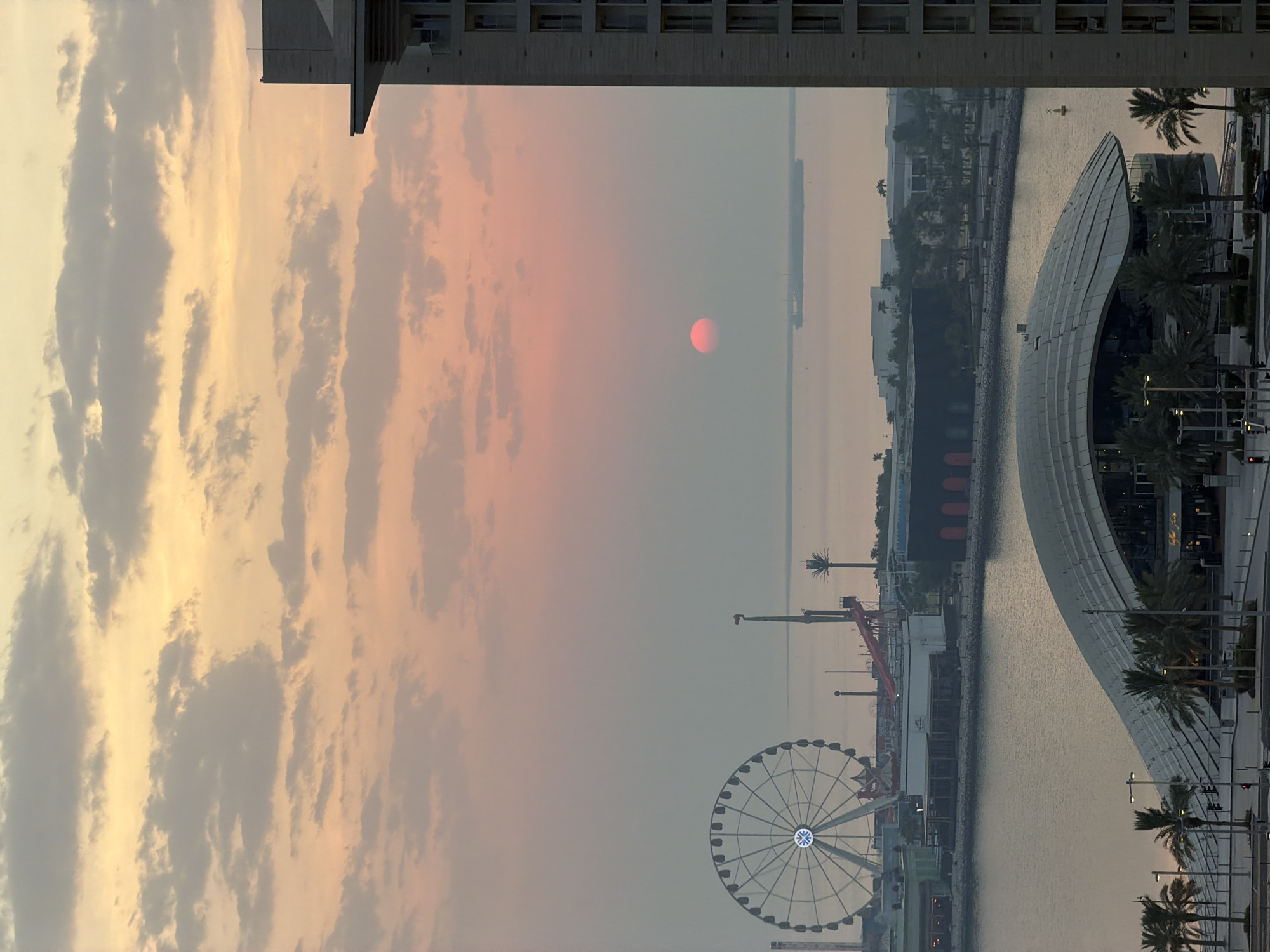 The image captures a sunset over the waterfront in Lusail, Qatar. A large Ferris wheel, illuminated with a blue light at its center, dominates the left side of the frame. Adjacent to it are various amusement park attractions, including a roller coaster track and a tall slender tower. In the foreground, a modern building with a distinctive curved roof, likely an entertainment venue or museum, is visible with palm trees lining its perimeter. A body of water separates the foreground from the amusement park area. Further out on the water, a cargo ship can be seen in the distance. The sky is filled with hues of orange and pink, indicating the sun is low on the horizon, partially obscured by a haze. A tall modern building, likely a residential or commercial tower, is positioned on the right side of the image, partially framing the scene. The overall atmosphere is serene and picturesque, typical of a coastal sunset.