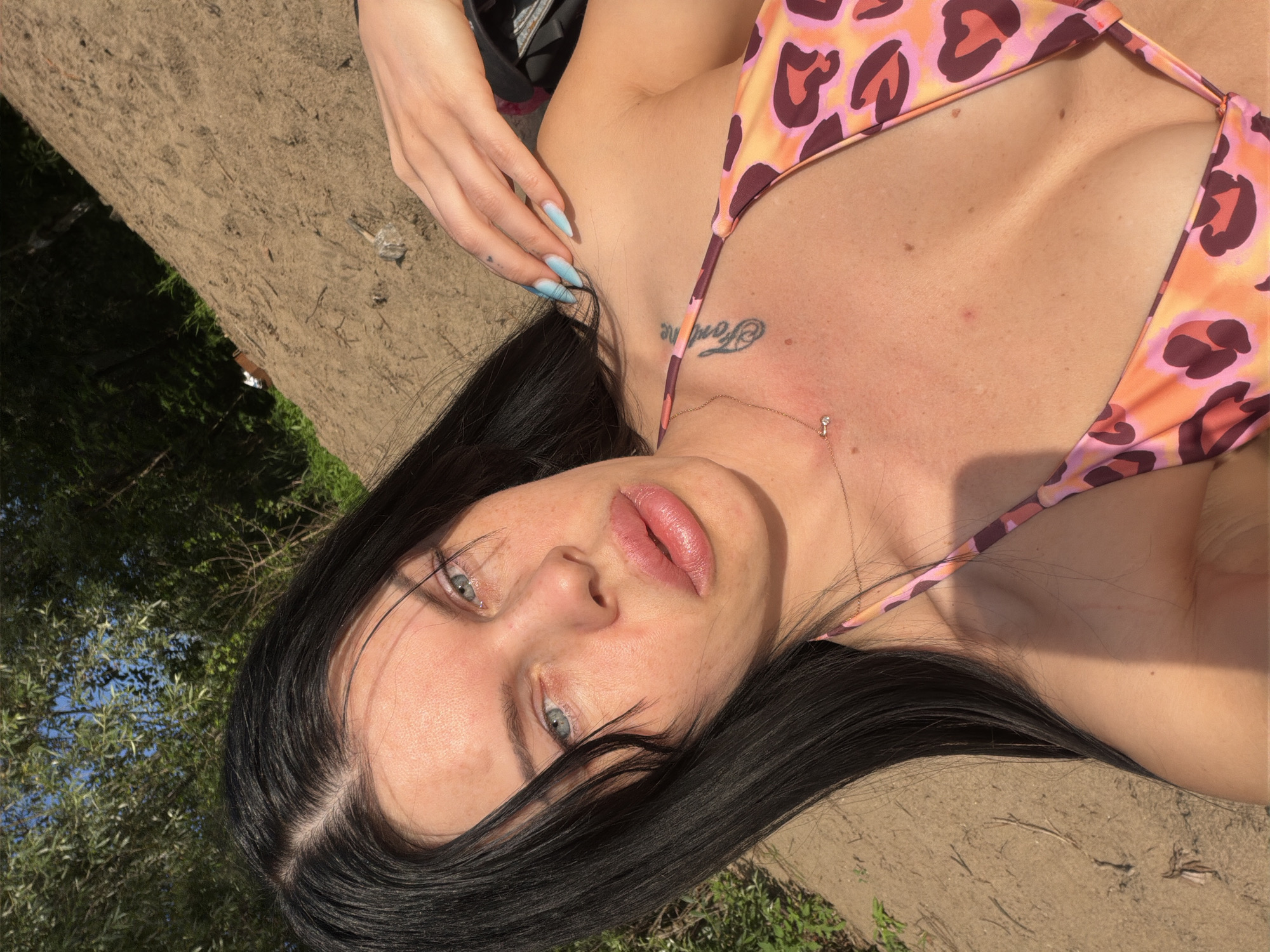 The media file shows a close-up, rotated shot of a young woman lying down outdoors. She has long, dark hair that falls around her face, which is looking upwards. Her eyes are a light blue-green, and she has full, pink lips. She is wearing a leopard-print bikini with pink and orange tones. A thin gold necklace with a small, clear pendant is visible around her neck. A tattoo is visible on her collarbone. Her left hand, with light blue-painted nails, is resting on the ground to her left.
The ground appears to be sandy or dirt, with some dry vegetation and twigs. In the background, out of focus, are green trees and a clear blue sky, suggesting it is daytime and the weather is fair. The sunlight casts shadows and highlights on her skin. The overall impression is of a relaxed, leisurely moment outdoors, possibly at a beach or in a park.
Visible text: There is a word tattooed on the woman's collarbone, but it is difficult to decipher clearly from the image.