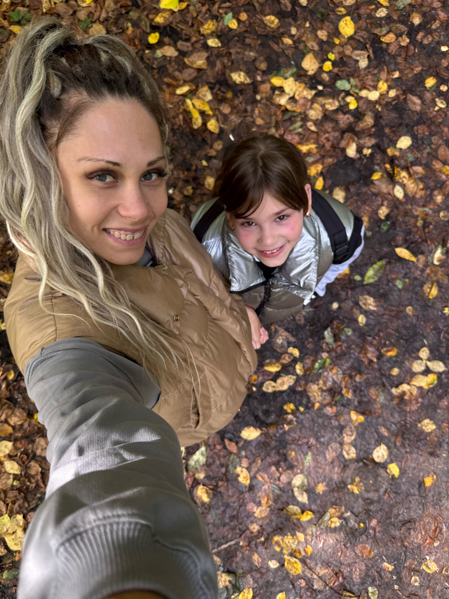 In Rusanovo, Russia, a woman with blonde hair in a ponytail and a tan puffer jacket is taking a selfie with a young person wearing a silver puffer jacket. They are standing on a path covered in fallen leaves, with the ground appearing damp. The woman is smiling at the camera, and the child is looking up and also smiling. The lighting suggests it is daytime, possibly with overcast skies as indicated by the soft shadows. The environment is a natural outdoor setting, likely a park or wooded area, with a focus on the autumn foliage on the ground.