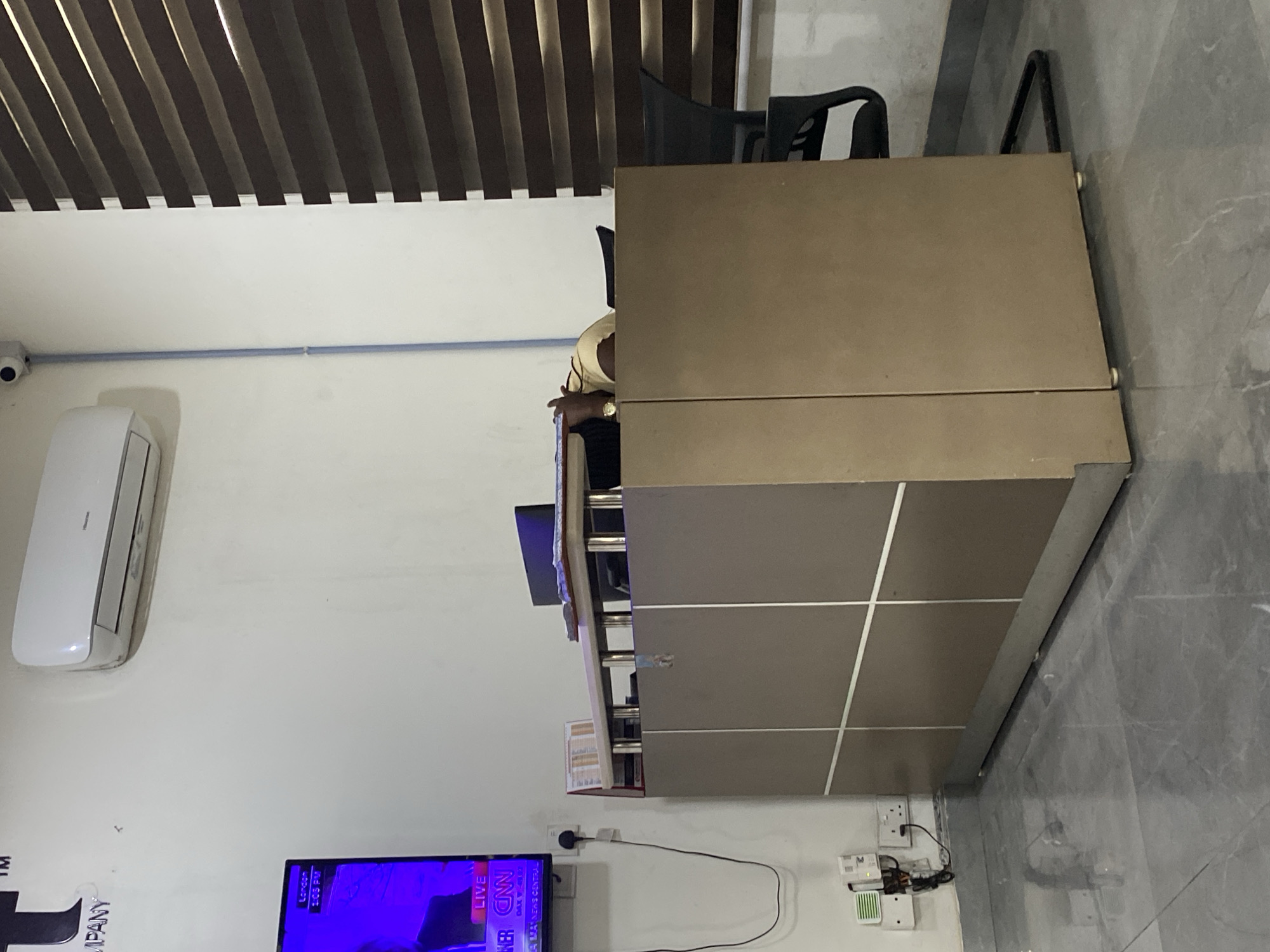 The image shows an indoor scene in what appears to be an office or commercial space. A light brown reception desk dominates the right side of the frame, featuring a segmented front with white dividing lines. Behind the desk, there is an office chair.
On the left, the wall is painted white and features an air conditioning unit installed high on the wall. Below it, a television screen displays content, with the CNN logo visible. Near the bottom of the wall, there are electrical outlets and related wiring. The ceiling has a brown slatted design. The flooring is a gray marble pattern.
It is impossible to ascertain the location as Ikeja, Nigeria from the image alone.
