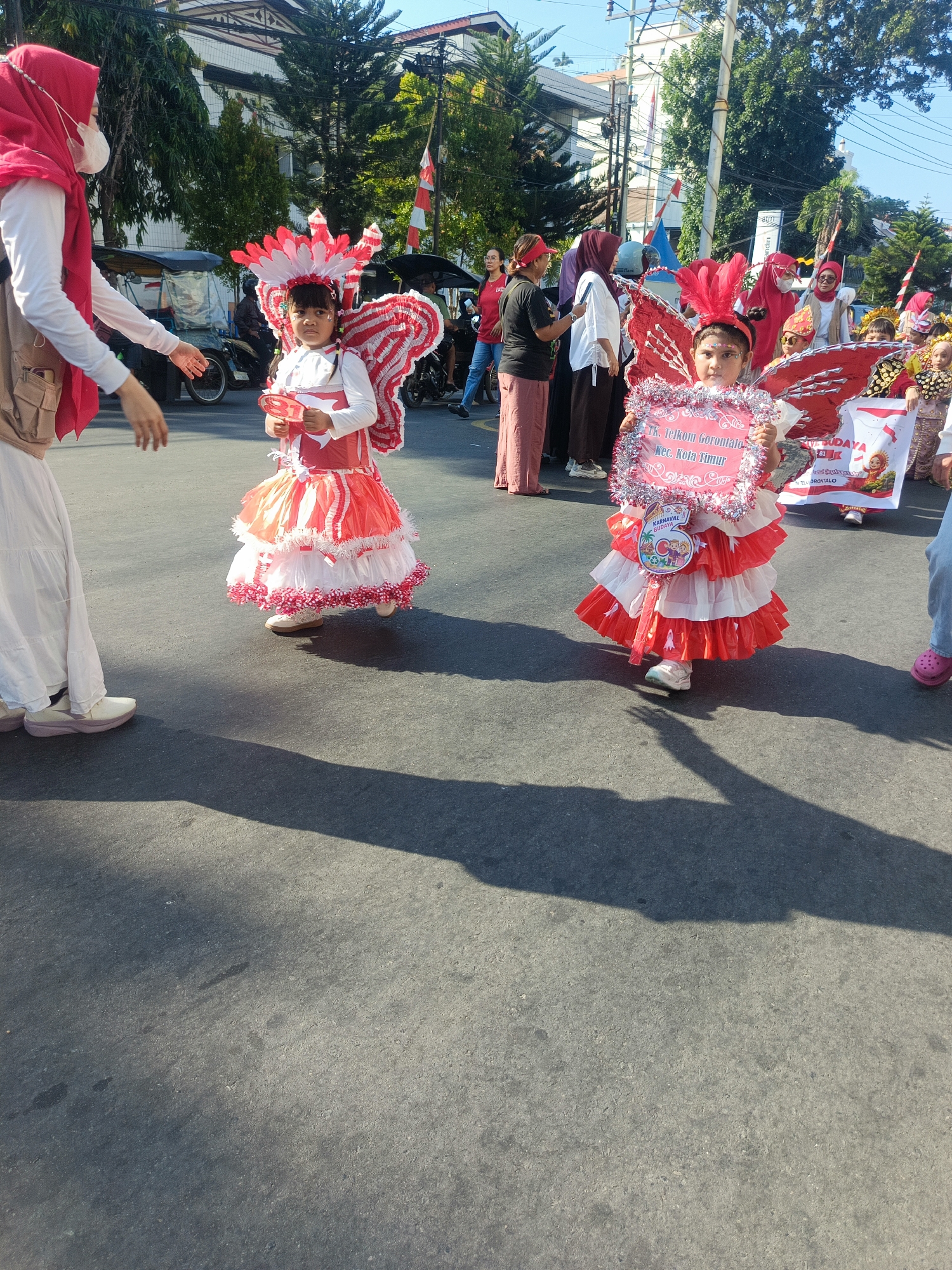 A bright outdoor daytime scene depicts a procession or parade on an asphalt road in Gorontalo, Indonesia. Two young girls, adorned in elaborate red and white costumes with butterfly-like wings and feathered headdresses, are the central figures. The girl on the right holds a decorated sign that reads "Tk. Telkom Gorontalo Kec. Kota Timur". Both girls wear white long-sleeved shirts beneath their costumes. A woman in a red hijab and white long-sleeved shirt is visible to the left, partially obscuring a tricycle in the background. Other individuals in festive attire and spectators are present in the background, along with trees and buildings. The sun casts long shadows across the road, indicating a sunny day.