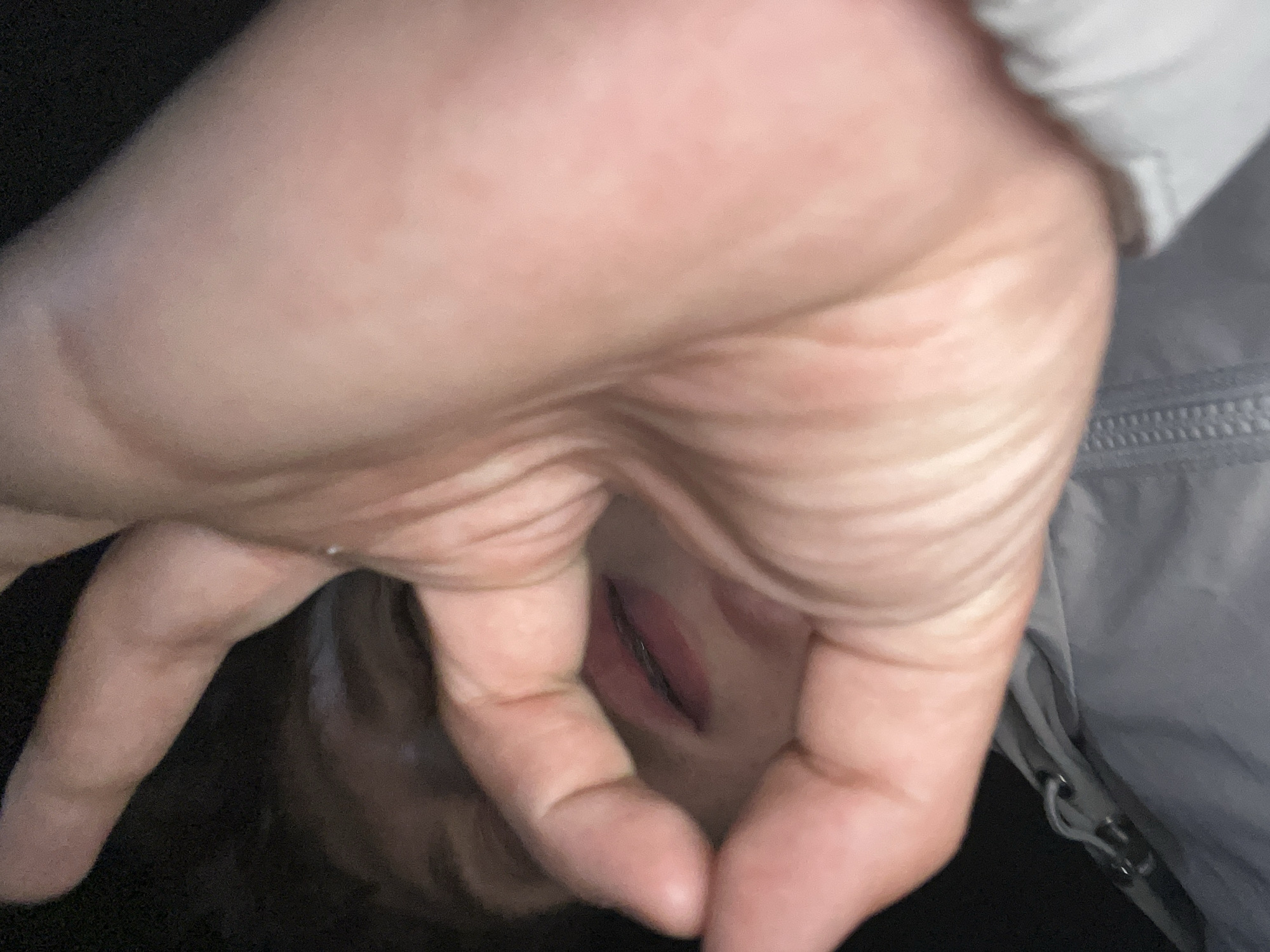 A close-up, low-angle, candid photograph captures a person's face mostly obscured by their hand, which is held up in front of their mouth, forming an "ok" sign. The person's lips are visible through the gap in their fingers, and they appear to be wearing lip gloss. The background is dark and out of focus, with hints of dark hair and a gray, zippered jacket. The image was taken at an unspecified outdoor location in Zlatograd, Bulgaria.
