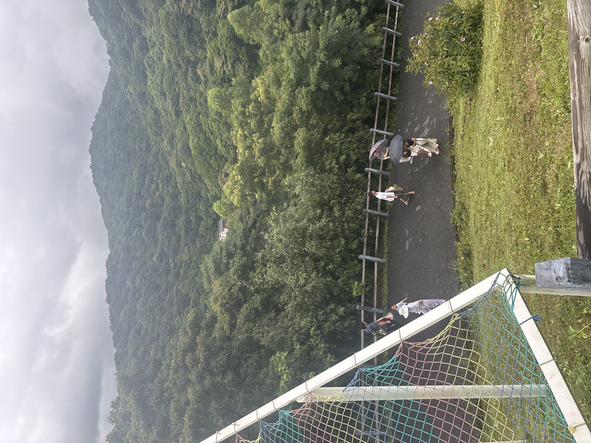 The image depicts a cloudy day with a lush, forested mountainside dominating the background. In the middle ground, a paved walkway runs along a wooden fence, with several people walking on it. A few individuals are using umbrellas, suggesting it may be raining or has recently rained. In the foreground, a section of a colorful net structure, possibly part of a playground or sports facility, is visible on the left. The ground slopes away from the walkway, covered in green grass. A small cluster of pink-flowered bushes is situated to the right of the walkway. The scene appears to be an outdoor recreational area in a hilly, natural setting. The location is not specified to be in Guangzhou City, China.