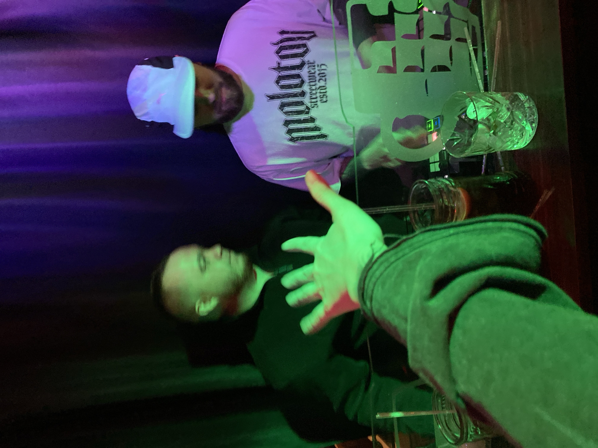 The image shows two men at a bar in Izhevsk, Russia. One man is wearing a white baseball cap and a light pink t-shirt with the word "Molotov" written on it. The other man, with a close-cropped haircut, is wearing a black shirt. A person wearing a green hoodie is reaching toward the table. There are two drinks with straws on a glass-topped table with a design etched into the glass. The lighting is dark and appears to be from colored neon or LED lights, with purple and green hues dominating the scene.