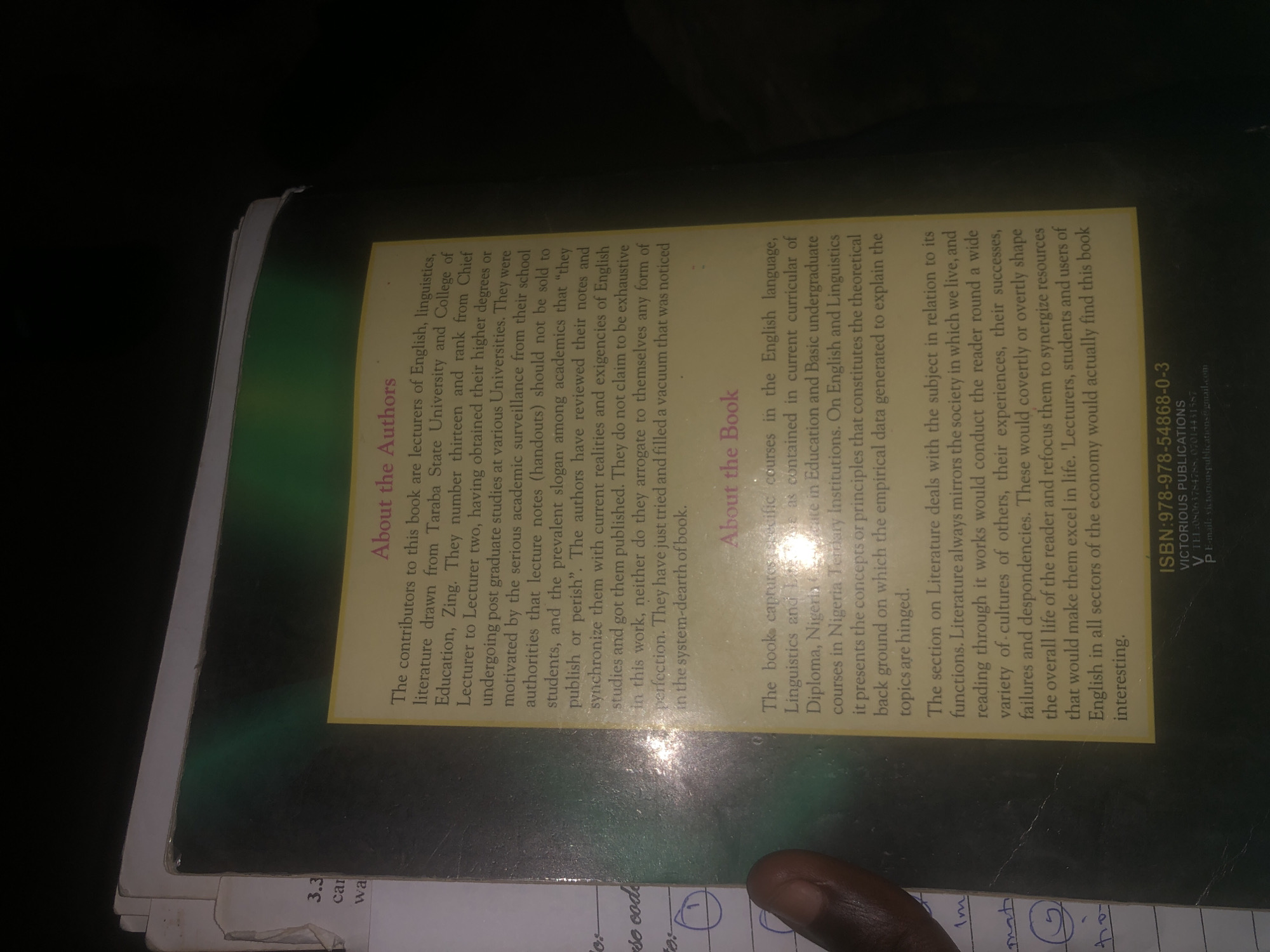 The image shows a close-up of the back cover of a book, predominantly featuring text. The book cover is a light yellow color with dark green and black accents. The text is black and pink. At the top of the visible text is a heading "About the Authors" in pink. Below this, a paragraph describes the contributors to the book. Further down, there is another pink heading "About the Book," followed by a description of its contents. At the bottom of the cover, there is a section with an ISBN number and publisher information.
To the left of the book cover, there are parts of other papers visible, with some handwritten notations including numbers and what appear to be circles.
The scene is indoors, likely a library or a study space, given the presence of books and papers. The lighting is dim, casting some glare on the book cover. There is no discernible location in Jalingo, Nigeria within the image.