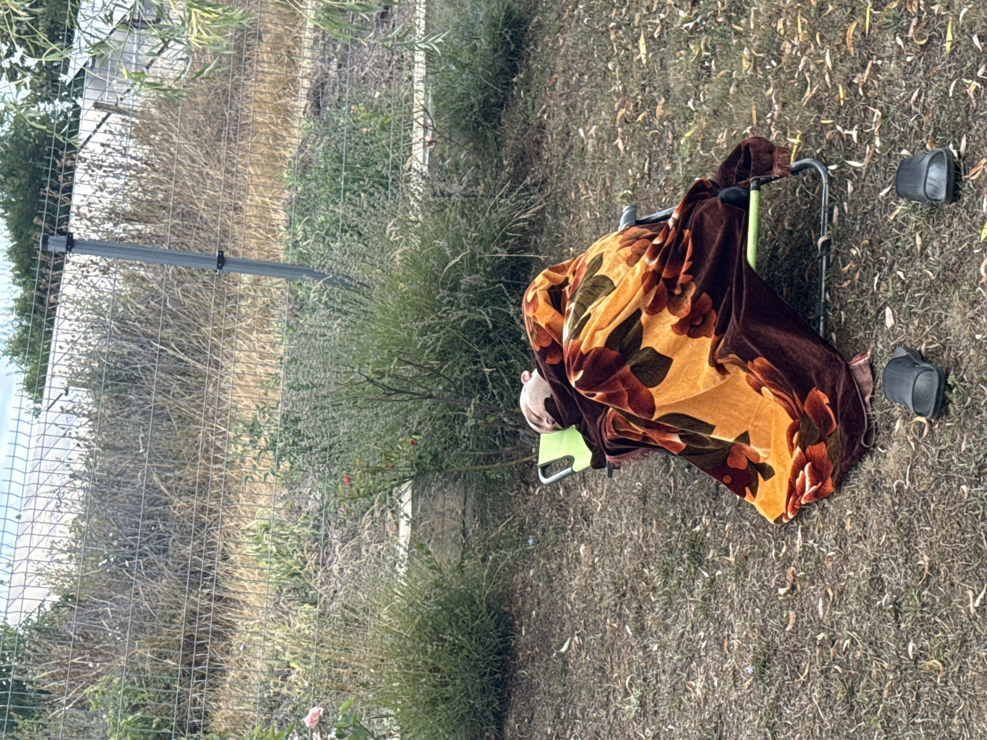 The image shows a person sleeping on a green chair outdoors, covered by a brown blanket with an orange and red floral pattern. Two black sandals are placed on the ground near the chair. The ground is covered in dry grass and fallen leaves. In the background, there is a wire fence and overgrown vegetation. The scene appears to be in a yard or garden area. The city of Vizitsa, Bulgaria, is not visually identifiable within the image.