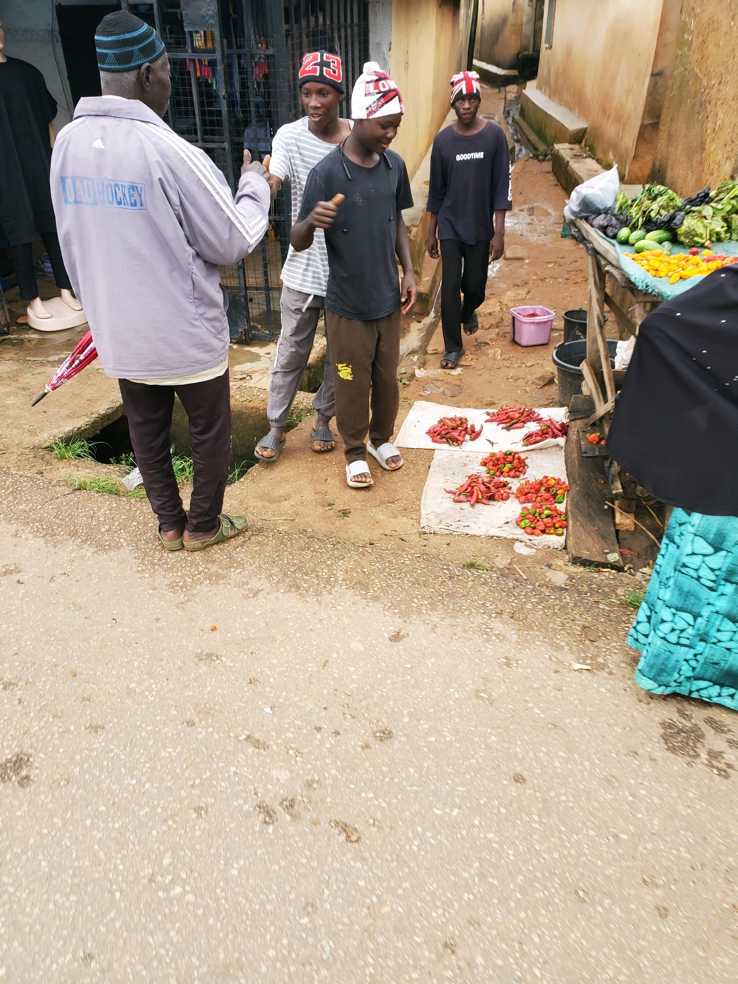 An outdoor market scene is captured in Jos, Nigeria, with a focus on a group of individuals interacting near a stall. The ground is a mixture of asphalt and dirt, with scattered debris and puddles, indicating recent rain.
In the foreground, an older man wearing a purple jacket with "DAD HOCKEY" printed on the back and a blue and black cap stands facing away from the camera, gesturing with his right hand. To his right, two younger individuals are visible. One wears a black t-shirt with "GOODTIME" printed on it and a red, white, and blue patterned beanie. He is looking towards the older man. Another young person, wearing a striped t-shirt and a red beanie with the number "23" on it, is also looking towards the older man and appears to be speaking.
Behind them, a wooden market stall displays a variety of fresh produce, including green vegetables, cucumbers, and piles of red and yellow peppers. A black fabric is draped over part of the stall, and a person in a turquoise patterned garment is partially visible to the right of the frame. Several buckets, including a pink one, are scattered around the stall area.
Further back, other figures are less distinct, suggesting a busy market environment. The architecture in the background consists of light-colored, weathered buildings. The overall atmosphere suggests a typical day at a local Nigerian market.