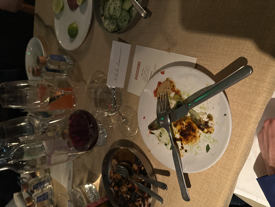 An overhead shot showcases a partially cleared table setting. Dirty white plates with used cutlery, including a fork and knife, suggest a meal has concluded. Several stemmed glasses, some containing remnants of red and orange liquids, are scattered across the surface. Small bottles labeled with blue markings lie among the glasses. A card with "Nikola Simic xxx" written on it sits near a menu labeled "DECIMO". Bowls of food scraps and assorted citrus slices complete the scene, set against a textured beige tablecloth.