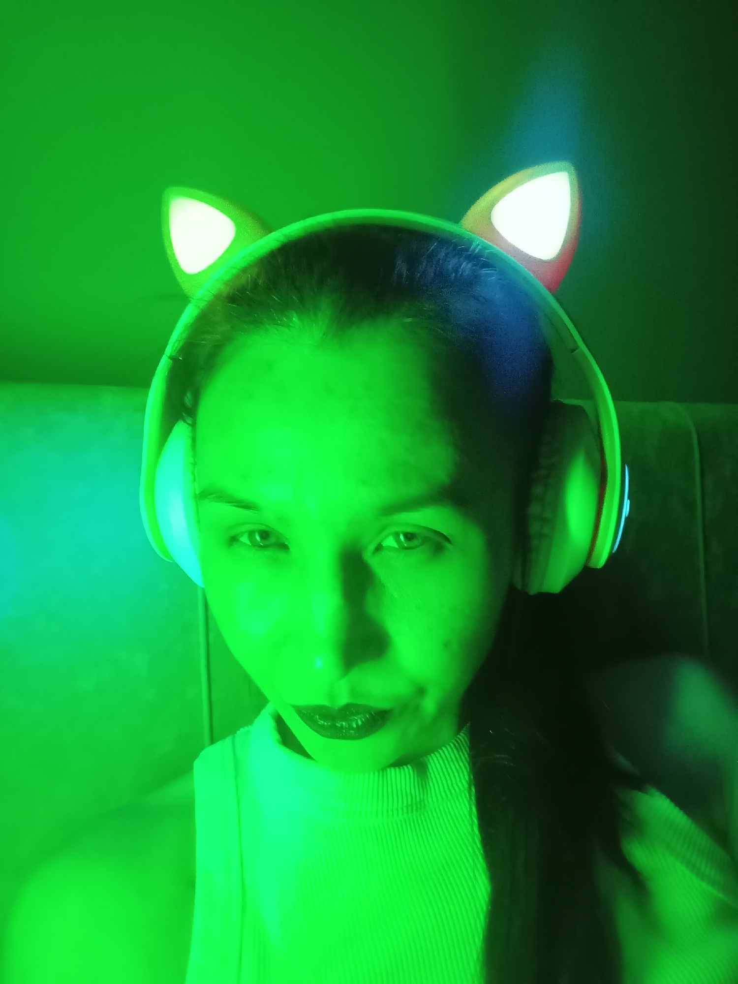 A head-and-shoulders portrait captured indoors in Pervomaysky, Russia, features a person, likely female, illuminated by a strong, vibrant green light. The intense green hue bathes the entire scene, including the subject's face, clothing, and the indistinct background, suggesting a low-light environment, possibly at night.
The person has dark hair and is wearing a light-colored, ribbed top. They are accessorized with over-ear headphones that have prominent, light-up cat ears on the headband. The cat ears glow white, with hints of reddish-orange trim, and a subtle blue light can be seen on the side of one headphone ear cup.
The subject is looking directly into the camera with a serious or neutral expression. Their lips are slightly parted, revealing what appear to be dental braces. The harsh green lighting creates dramatic highlights and shadows across their features. The background is a soft, dark green blur, consistent with an indoor setting. There is no visible text in the image.