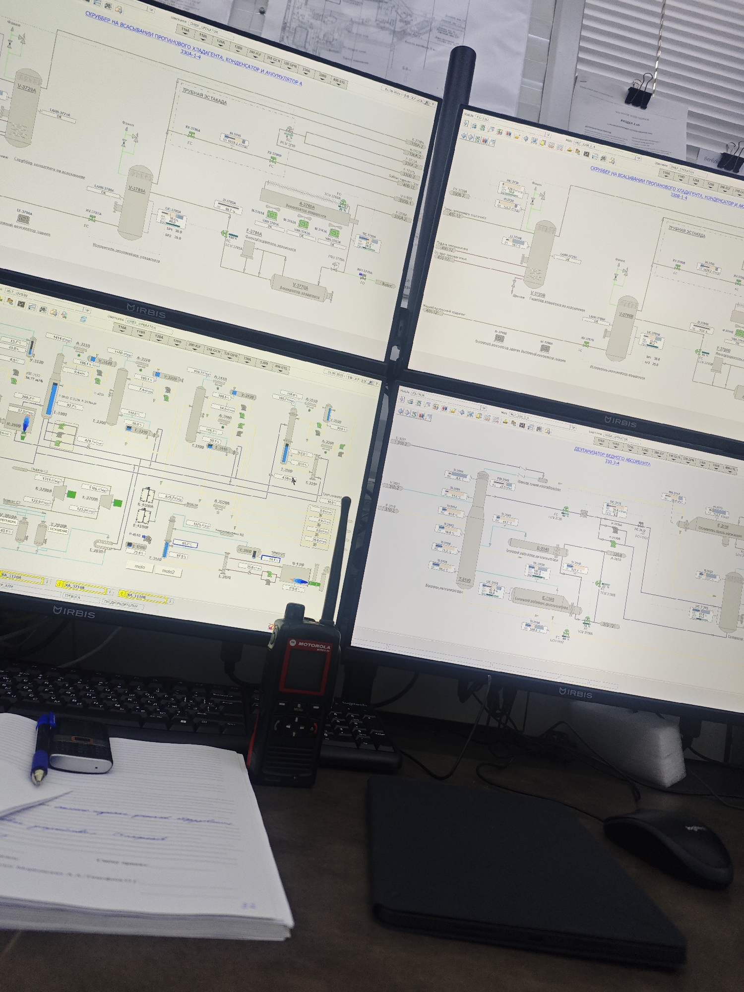 The scene appears to be a control room or workstation, focusing on four computer monitors displaying process flow diagrams, likely from an industrial plant. A Motorola walkie-talkie sits on the desk in front of the monitors, along with a keyboard, mouse, notepad, and pen. The diagrams show various equipment, including scrubbers, condensers, and accumulators, with numerical readouts and process indicators. The system seems to be monitoring and controlling the flow of propane refrigerant, as indicated by text on the screens. The environment suggests an industrial setting where real-time monitoring and control of chemical processes are essential.