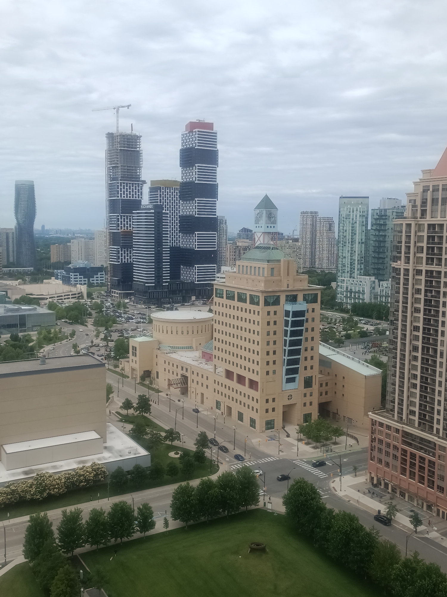 The image displays a wide-angle, high-angle view of a city skyline, specifically Mississauga, Canada. The scene is dominated by modern architecture, including numerous tall buildings, some of which are under construction. A prominent feature is the distinctively designed "Exchange" building, a cluster of towers with contrasting black and white facades, with one tower still being built with a crane visible. To the left, another tall, curved skyscraper is visible. In the foreground and mid-ground, a large, light-colored, multi-story building with many windows is present, featuring a unique blue and glass-paneled vertical element. Adjacent to it is a lower, circular structure. The street level shows vehicles navigating roads and a parking lot filled with cars. Green spaces, including manicured lawns and trees, are interspersed throughout the urban landscape, particularly in the lower portion of the image and around the buildings. The sky is overcast, suggesting a cloudy day.